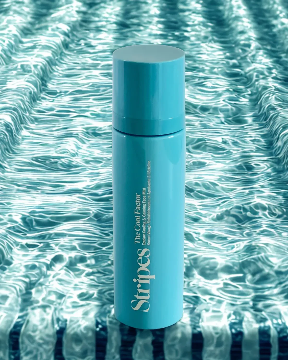 A blue bottle of face mist with the words 'The Cool Factor' on it, standing on a swimming pool deck with water ripples in the background.