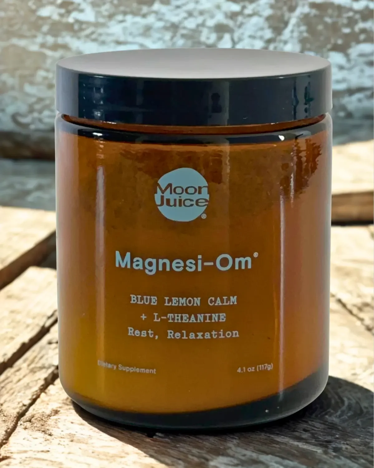 A brown container of Moon Juice Magnesi-Om supplement with a gray lid, placed on a wooden surface outdoors.