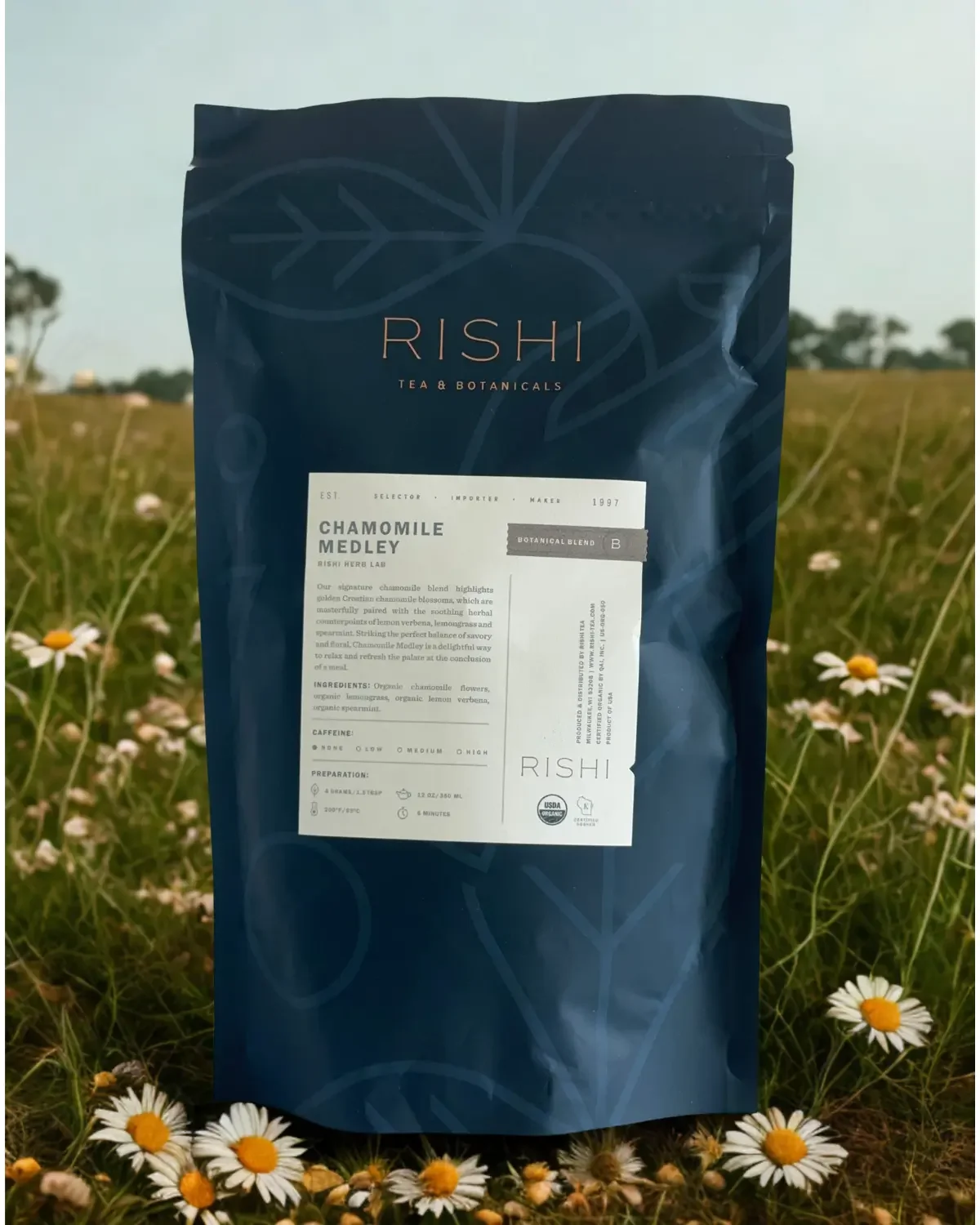 A blue package of Rishi Tea & Botanicals Chamomile Medley sitting in a field of daisies with a cloudy sky in the background.