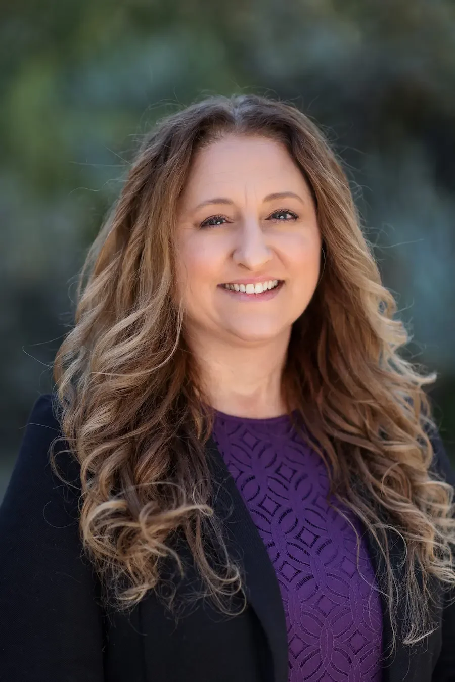 A woman with long, curly blonde hair, smiling with white teeth, wearing a dark blazer and purple patterned top, outdoors with blurred greenery background.