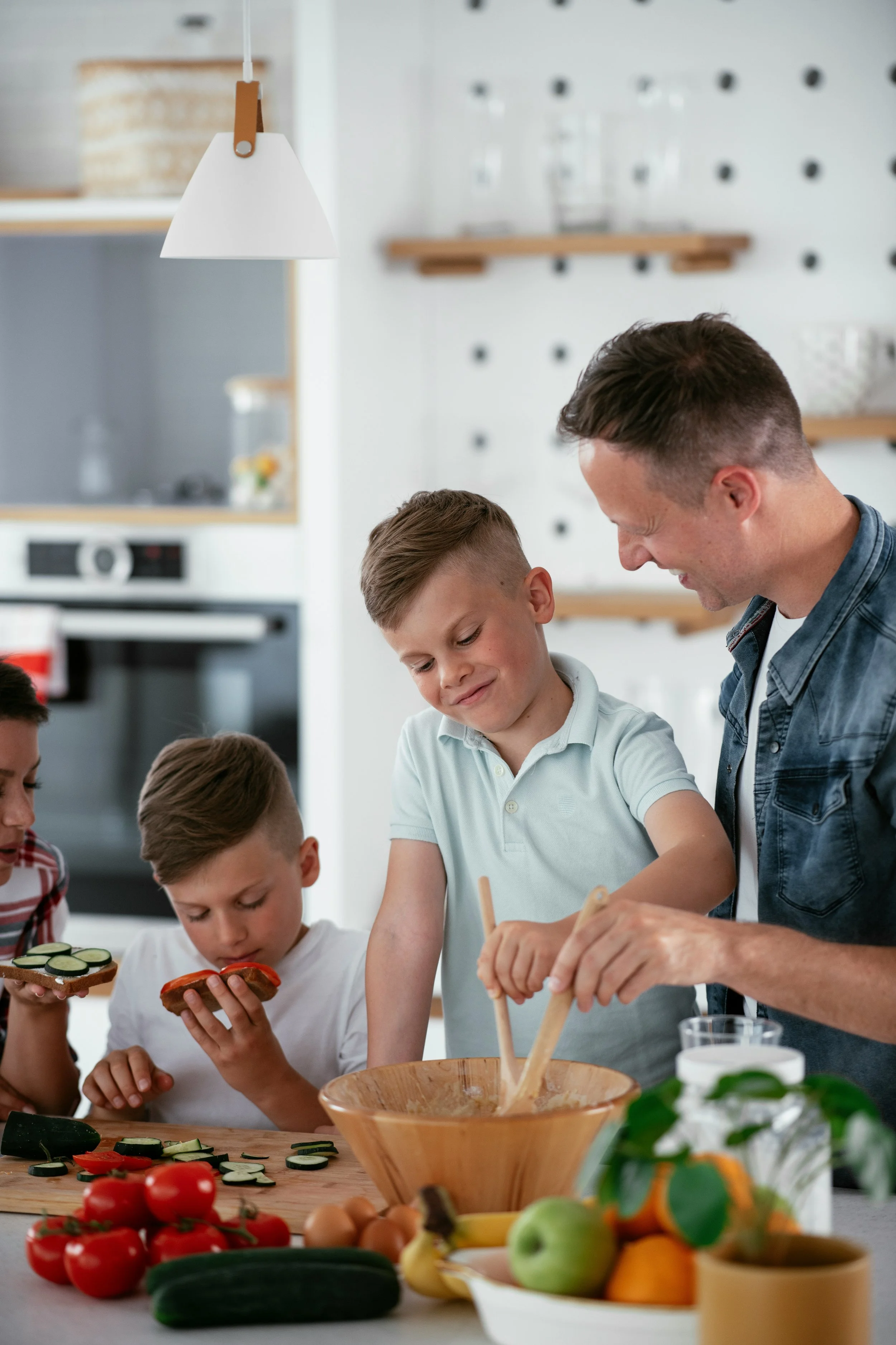 A man and three children preparing food together in a kitchen, with vegetables and bread on the table, and mixing in a large bowl.