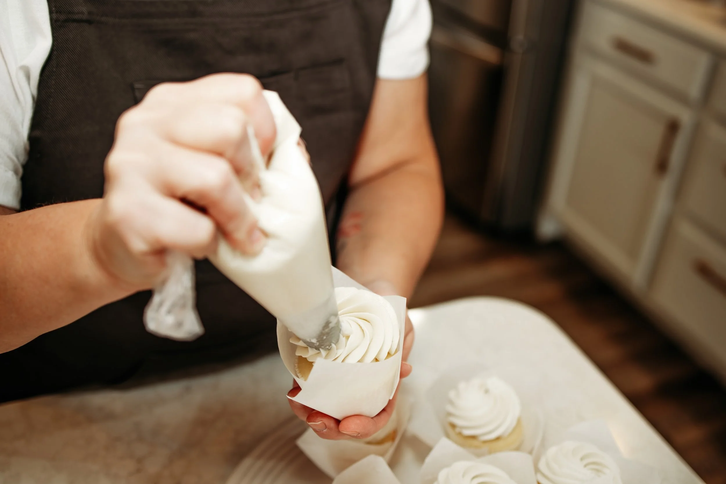 A person piping white frosting onto cupcakes in a kitchen.