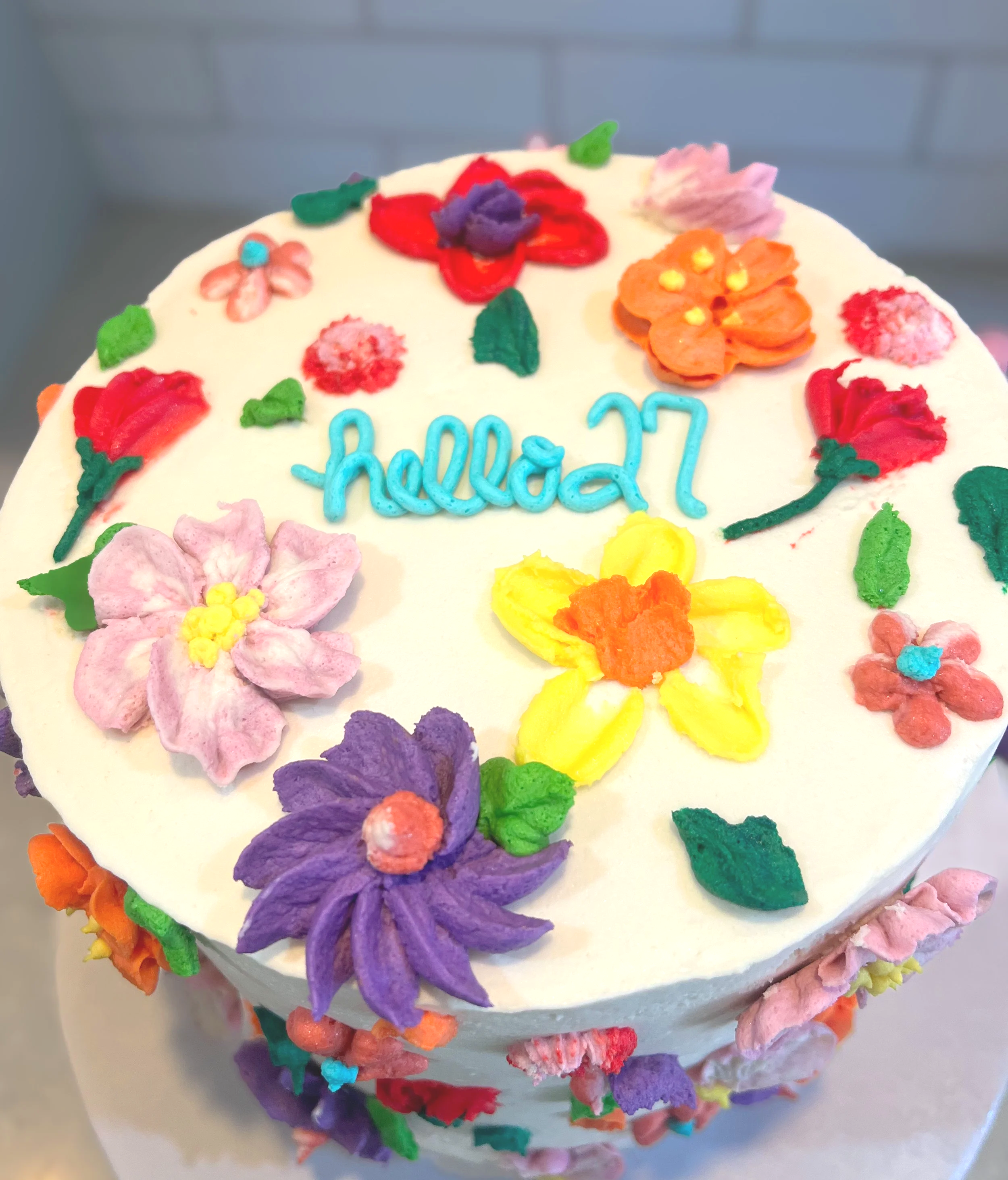 A round white birthday cake decorated with colorful fondant flowers and the word "hello" in blue icing.
