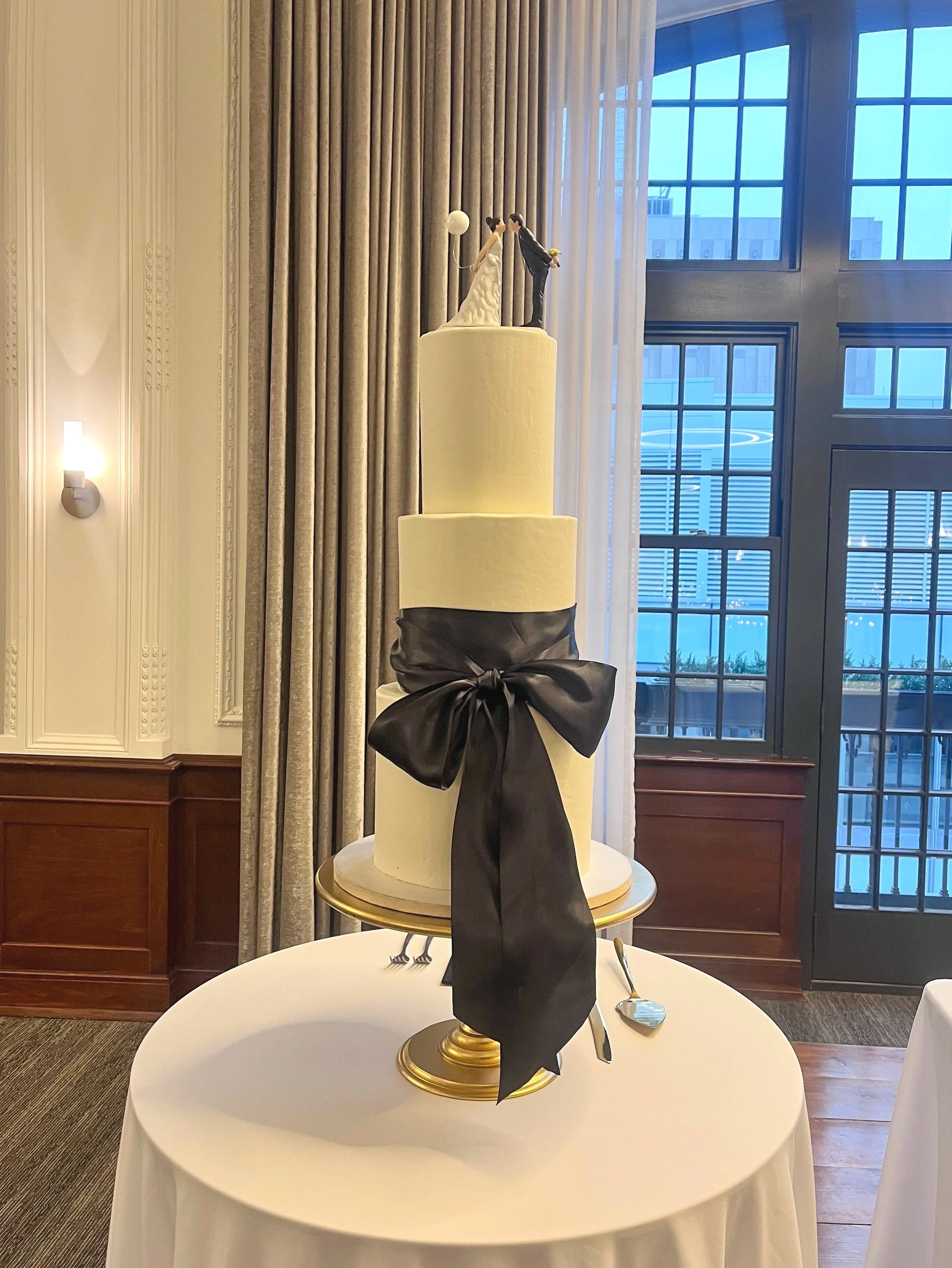 Elegant wedding cake with two tall white tiers, decorated with a black ribbon bow, topped with a figurine of a bride and groom in wedding attire, set on a round table with silverware. The background features large windows and curtains.