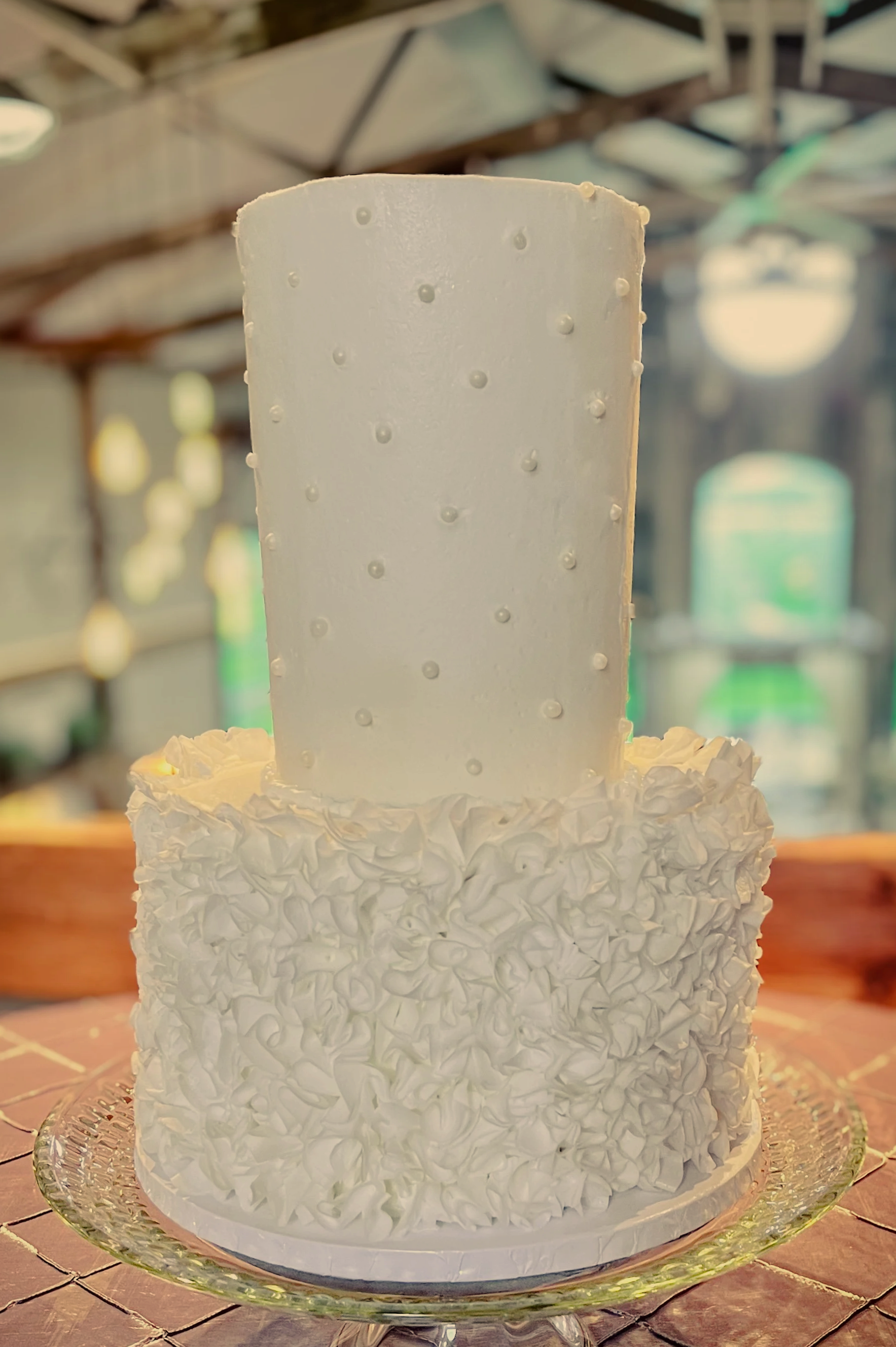White wedding cake with textured icing and a tall, smooth rectangular topper, displayed on a glass cake stand on a textured tablecloth, inside a rustic venue.
