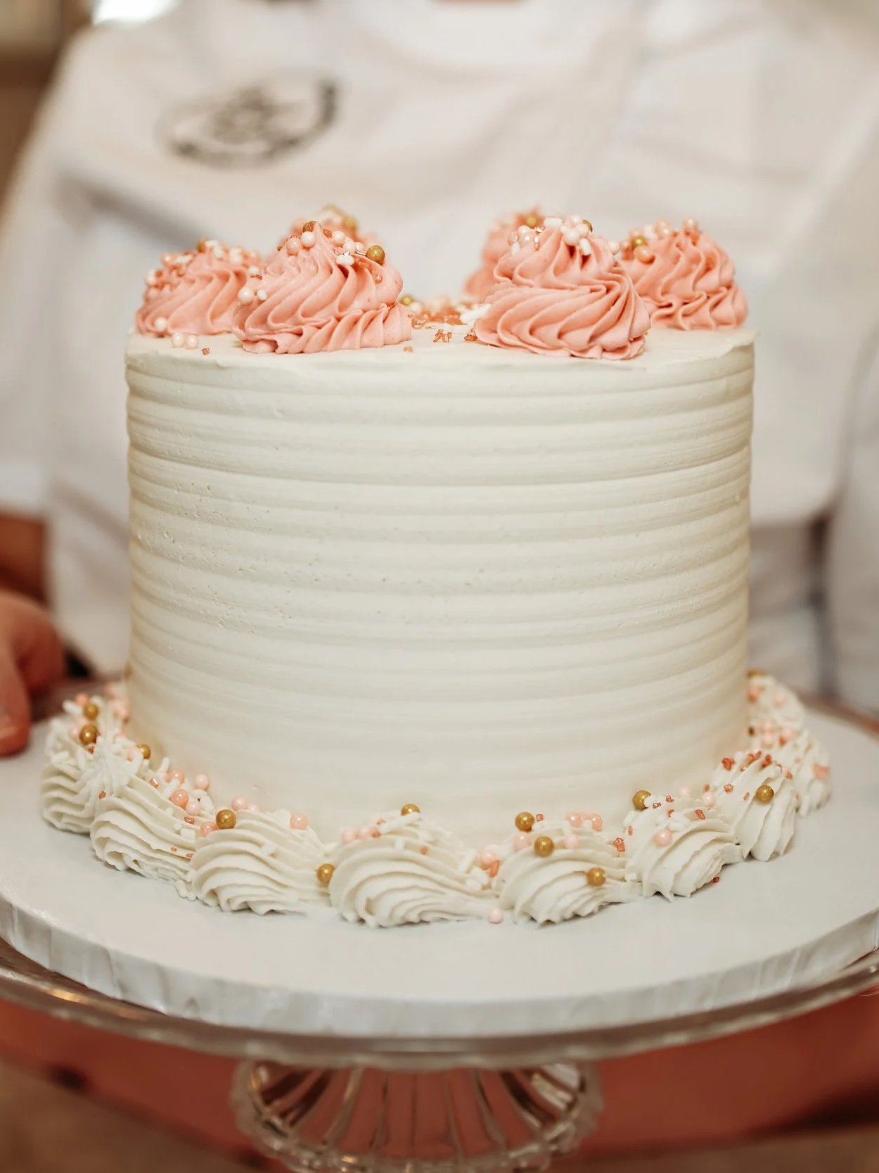 A person holding a white frosted cake with pink and gold decorative frosting on top and around the base.