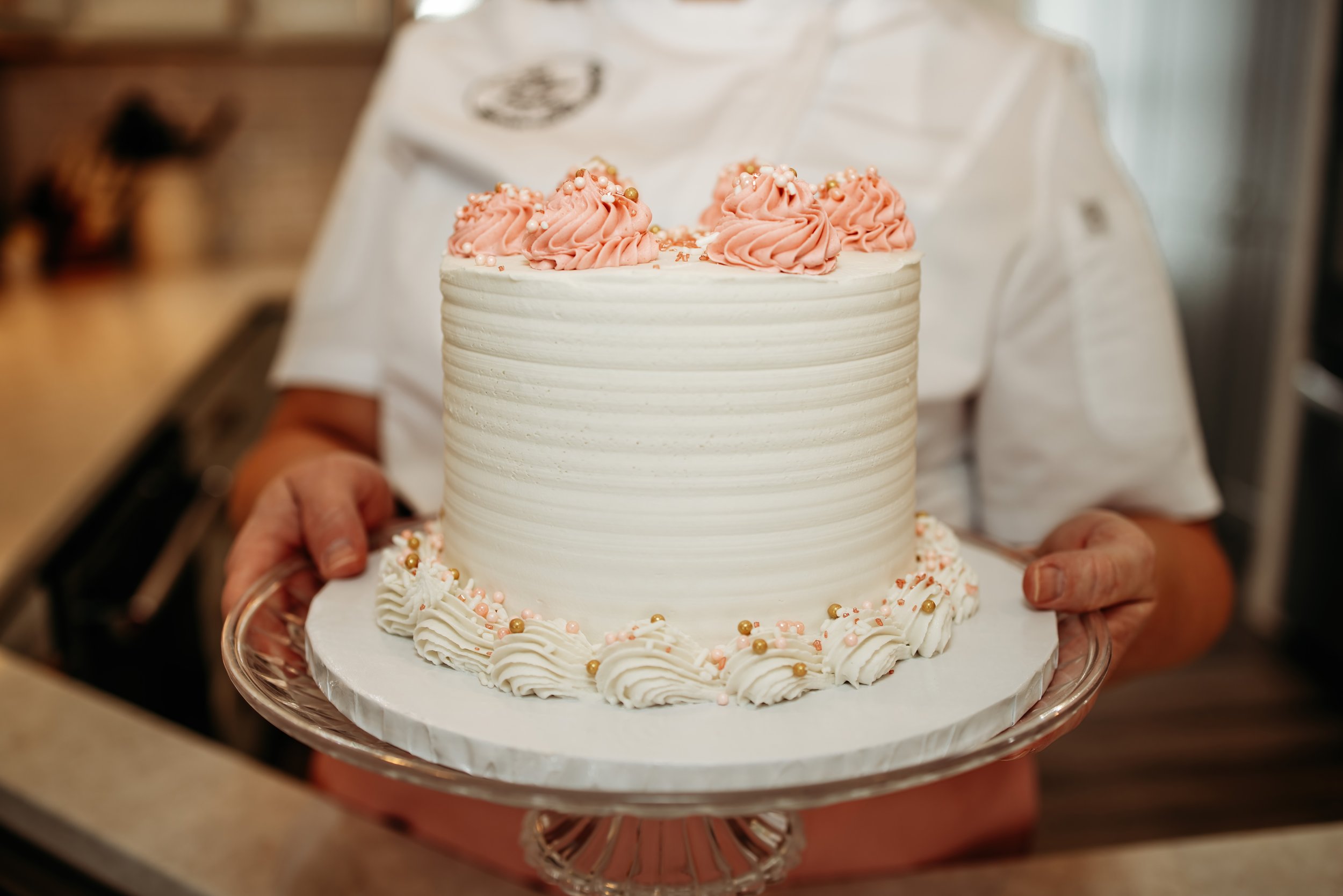 A person holding a white frosted cake with pink and gold decorative frosting on top and around the base.