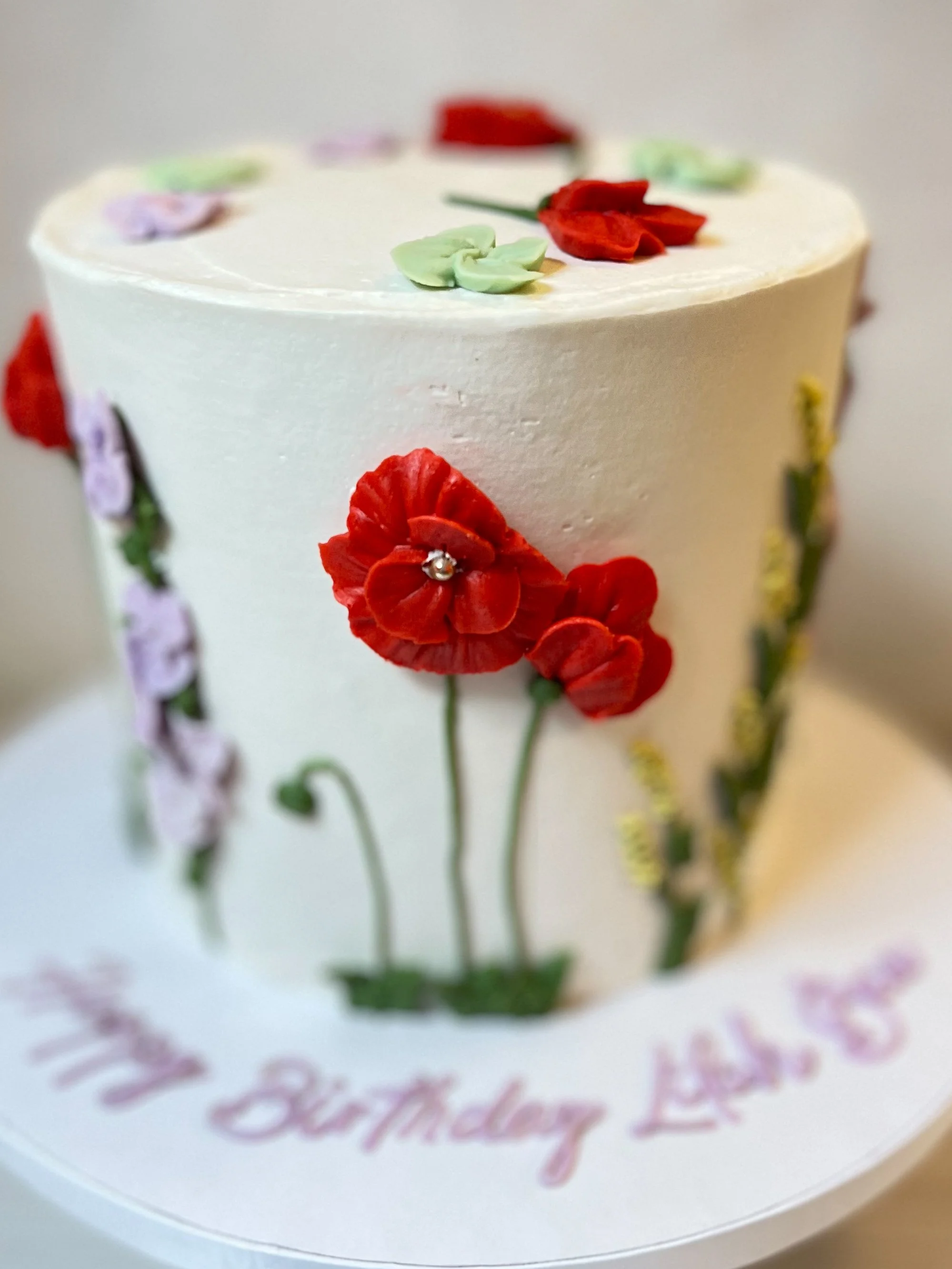 A white birthday cake decorated with red, purple, green, and yellow edible flower decorations, with a message for Lisa's birthday written in pink icing on the cake board.