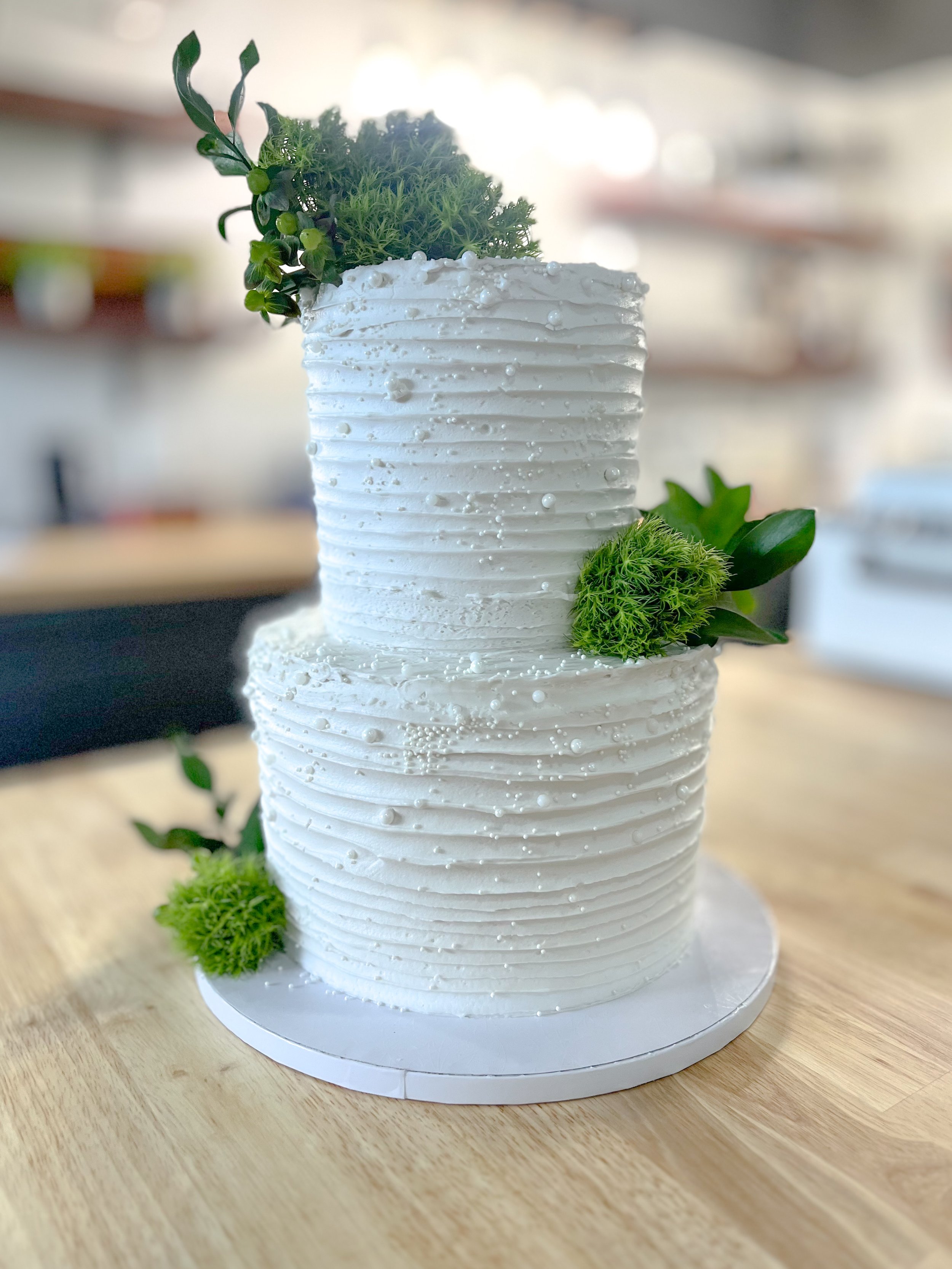 A two-tiered white wedding cake decorated with small green plants and white edible pearls, placed on a wooden table.