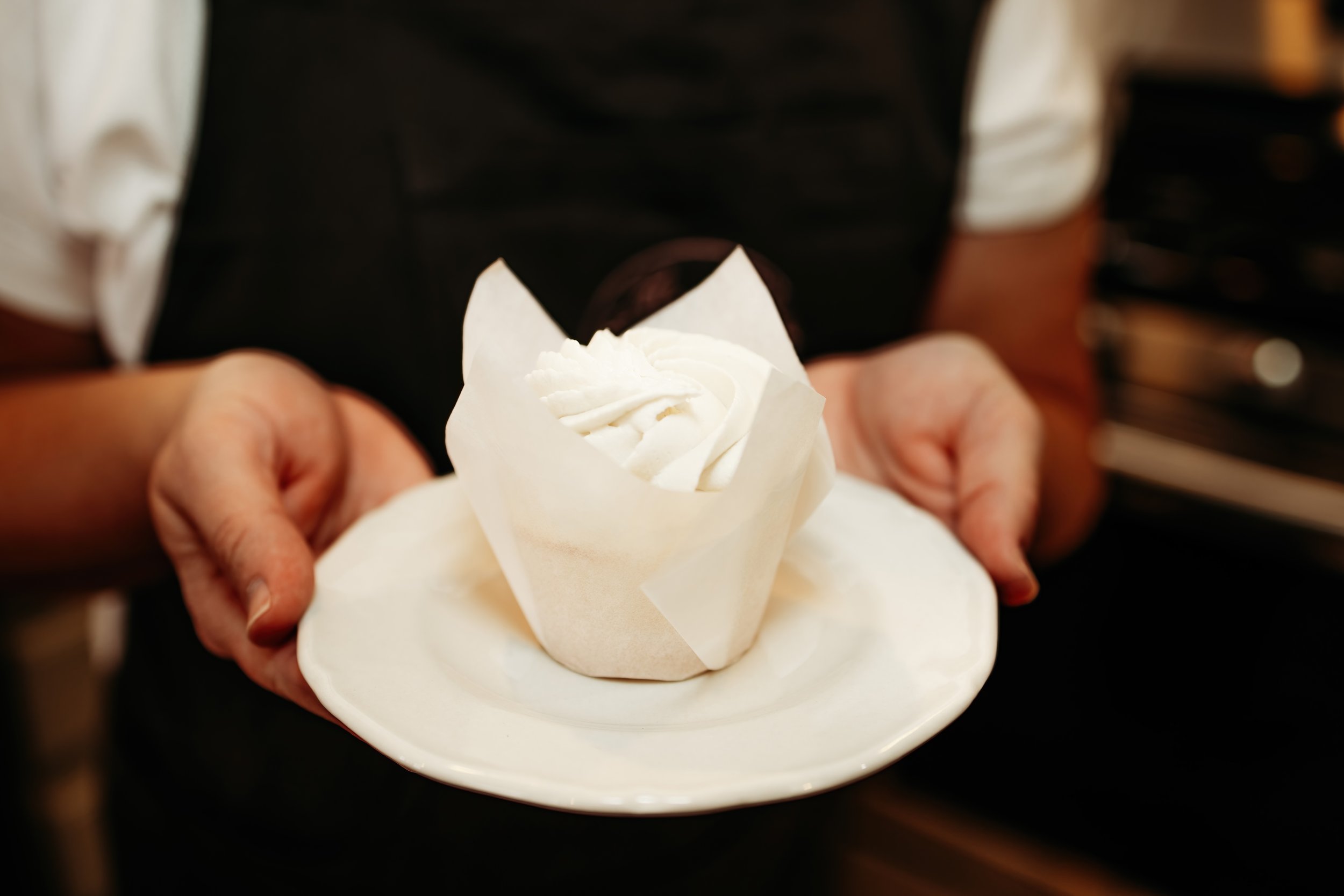 A person holding a white plate with a cupcake wrapped in parchment paper and topped with whipped cream.