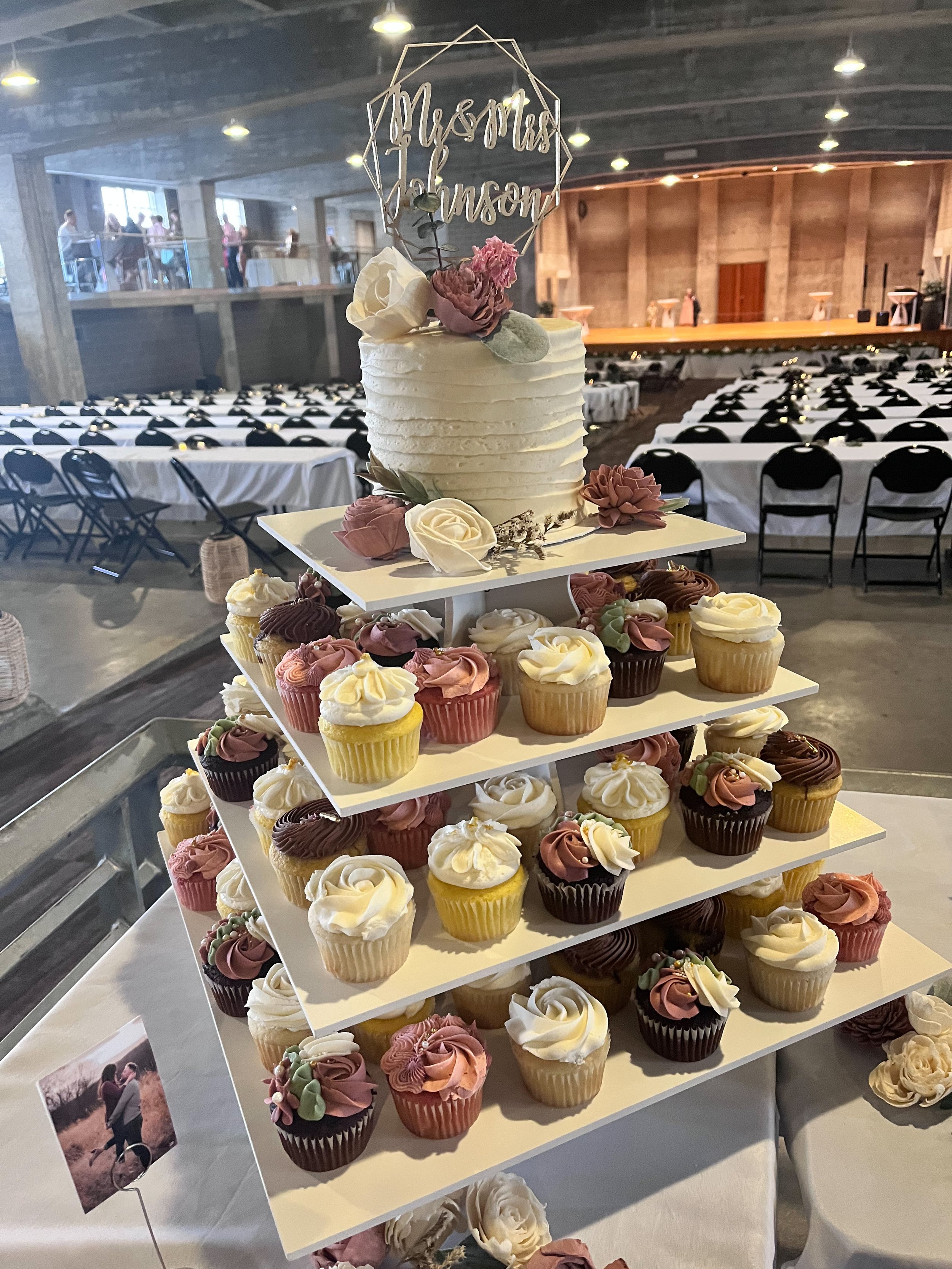 A wedding cake topper with the words 'Mrs. and Mr. Johnson' above a white frosted cake surrounded by colorful cupcakes on a tiered display in a decorated event hall.