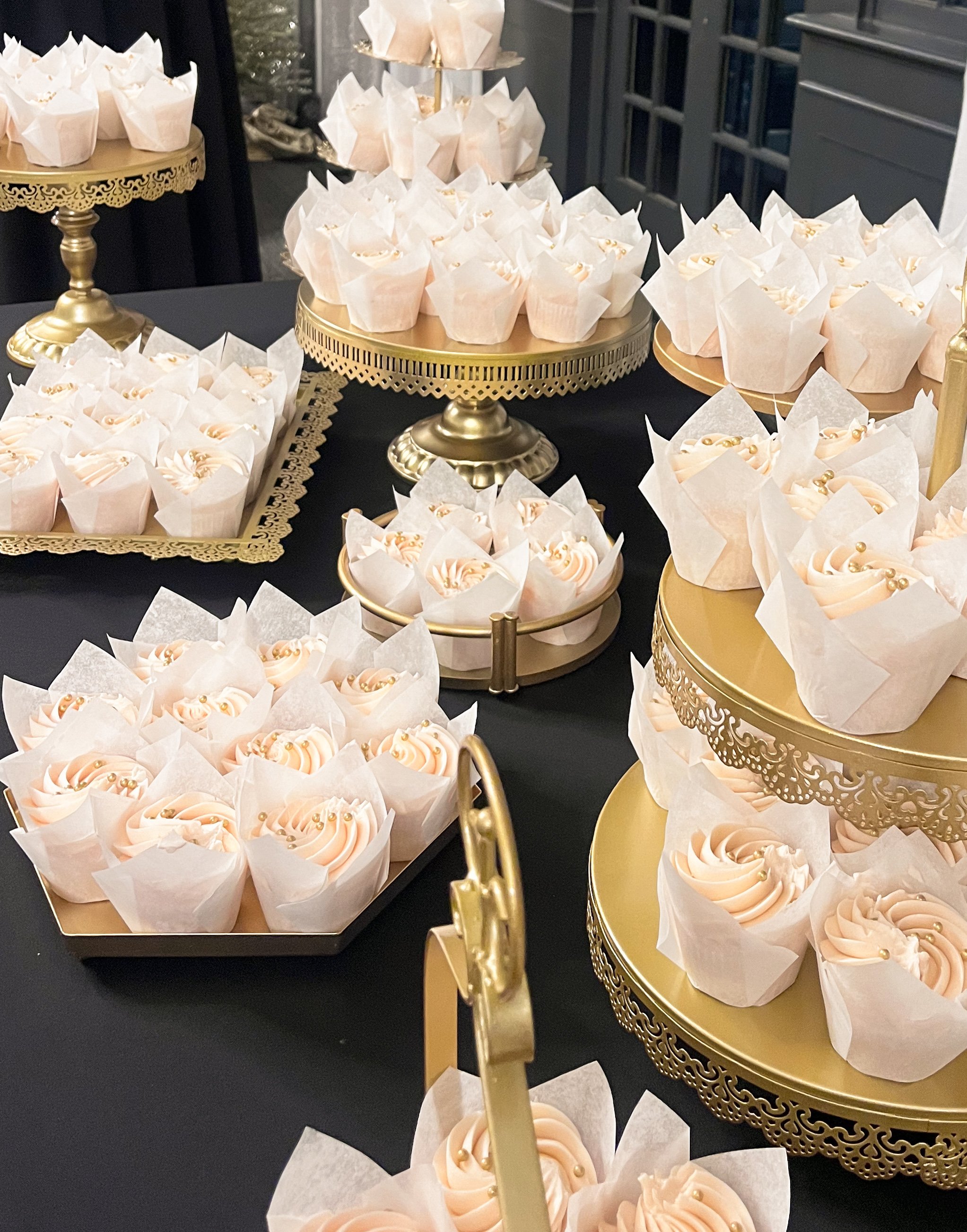 An array of elegant cupcakes with light pink frosting and gold pearl decorations, displayed on various gold and black tiered trays at a dessert table.
