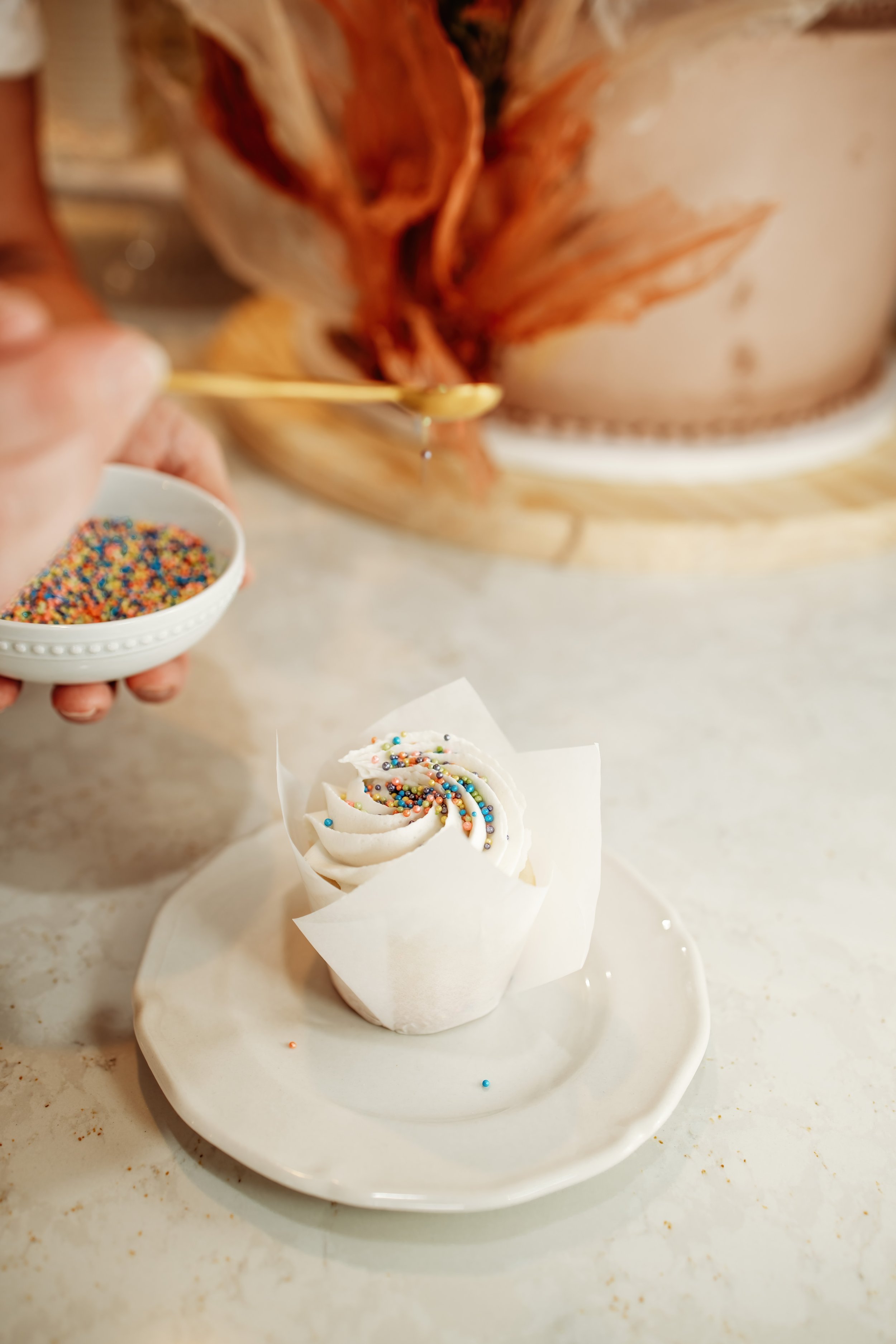 A cupcake with white frosting and multicolored sprinkles on a white plate. A hand holds a small white bowl of sprinkles, and another hand drizzles honey or syrup onto the cupcake.