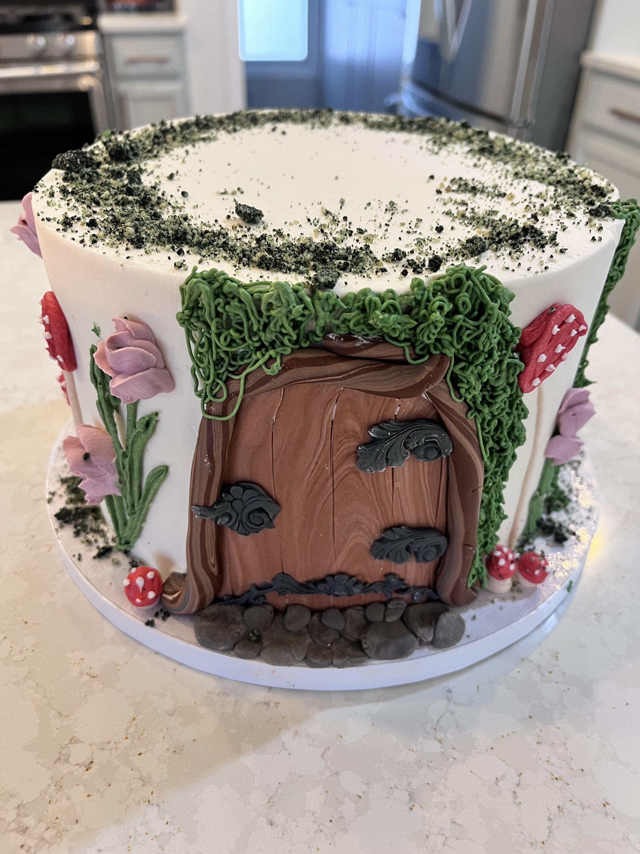 A decorated cake with a fairy tale door, surrounded by pink flowers, green frosting leaves, and mushrooms, topped with crushed cookie soil.