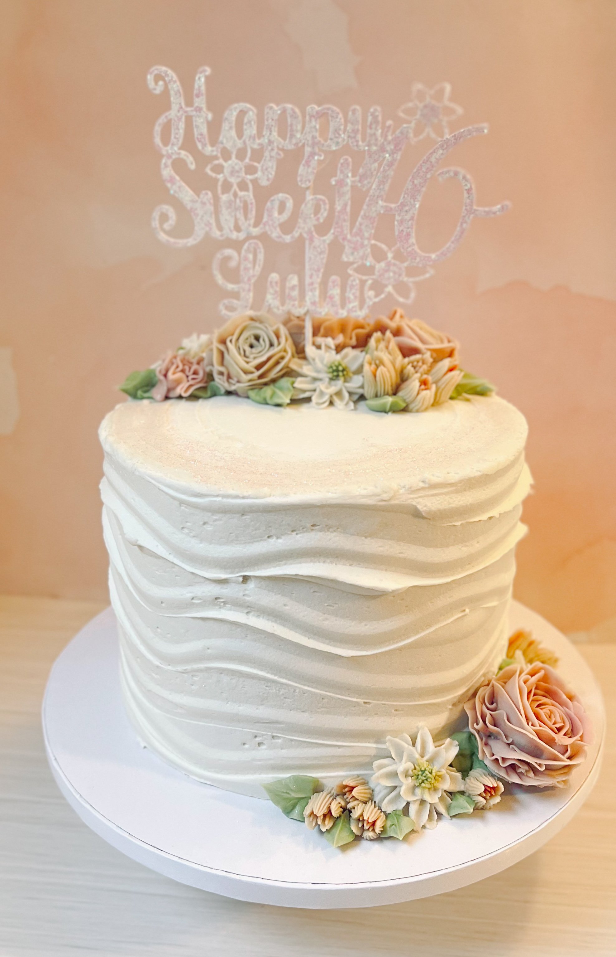 A white frosted birthday cake decorated with pastel-colored flowers and a glittery topper that says "Happy Sweet 16" with a star and flower design.