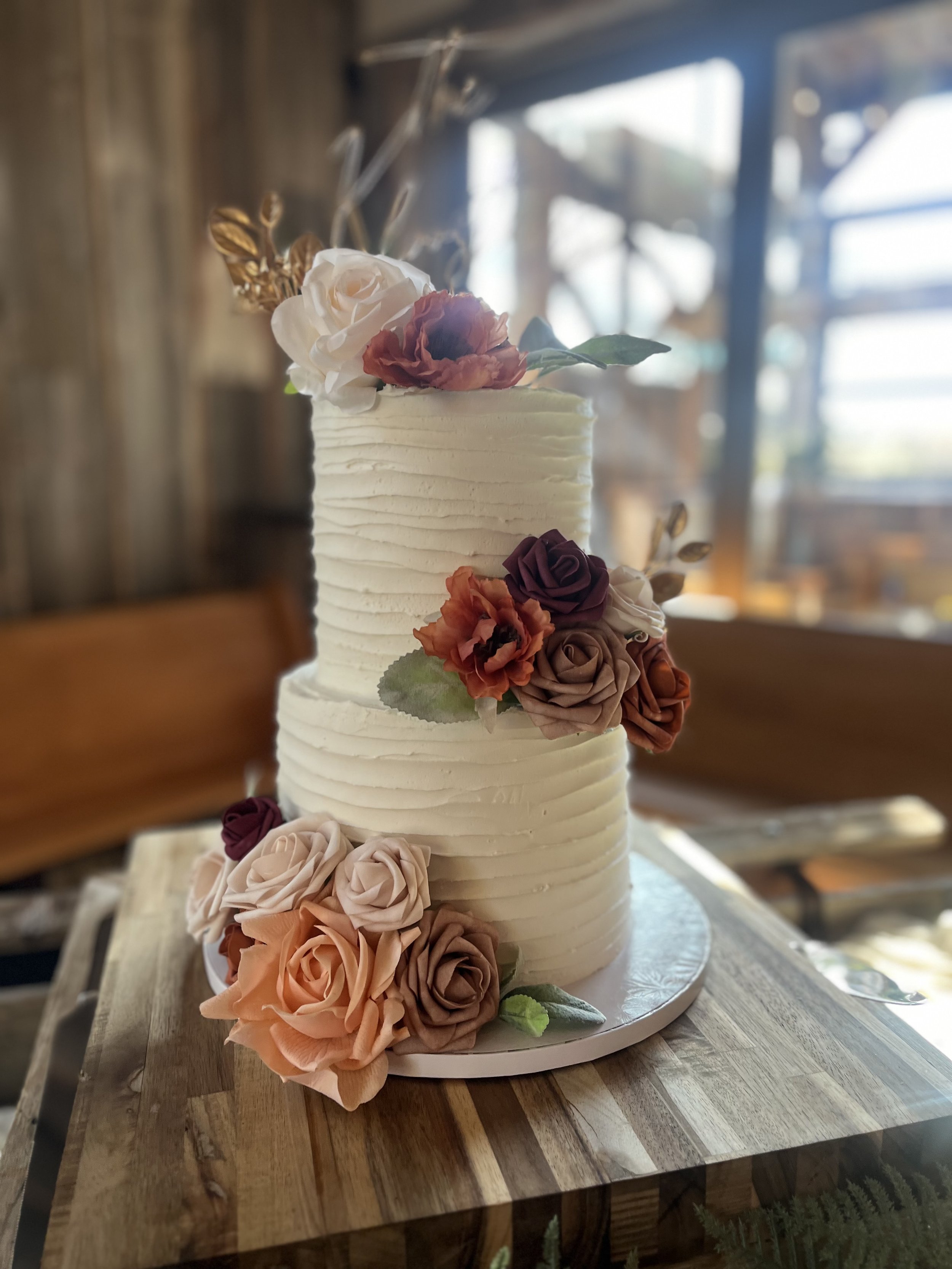 A two-tiered white wedding cake decorated with colorful sugar flowers, including roses and other blooms, placed on a wooden table inside a rustic venue with wooden walls and large windows.