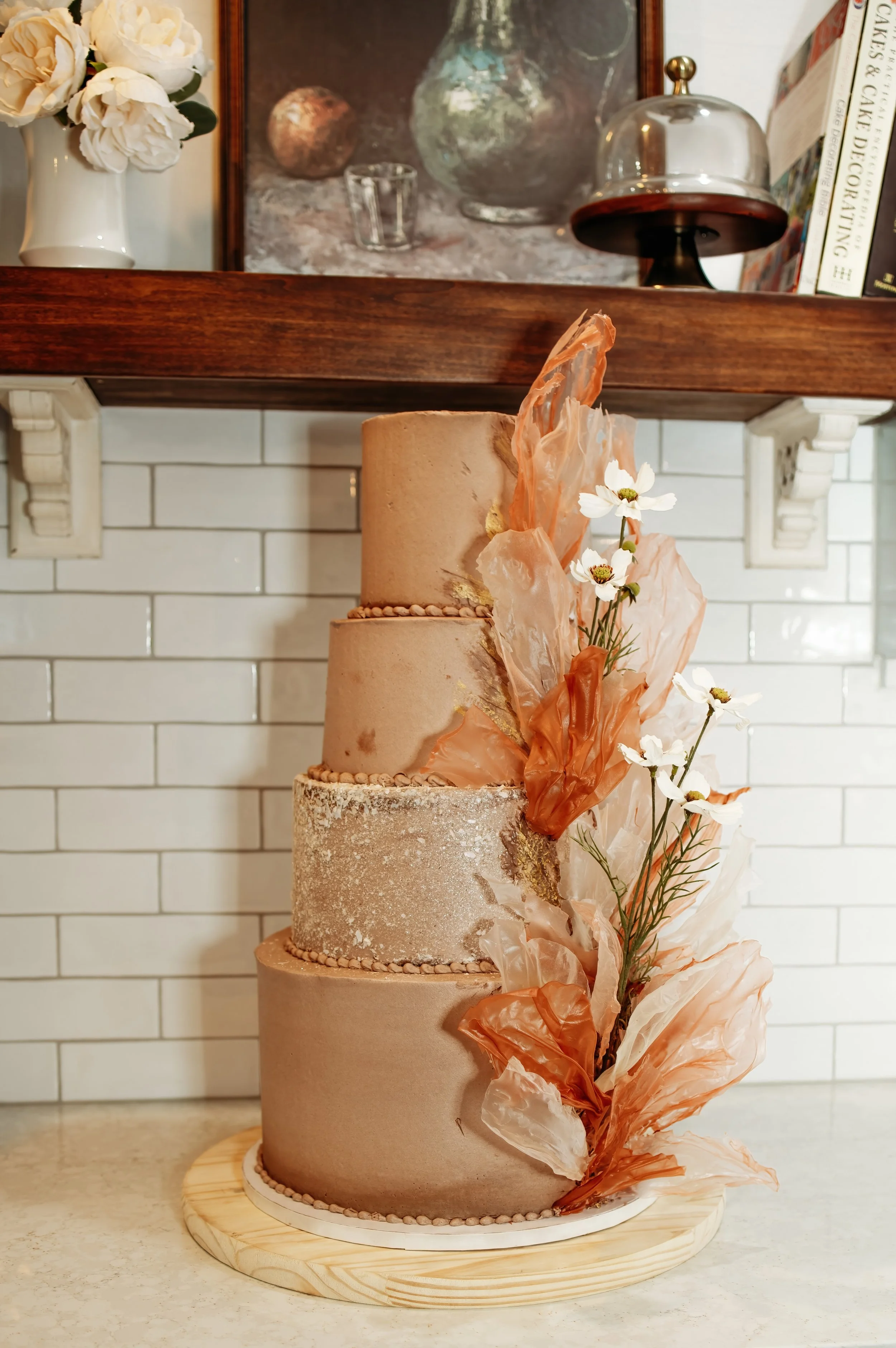A four-tiered beige cake decorated with edible gold accents and white flowers, placed on a wooden board.