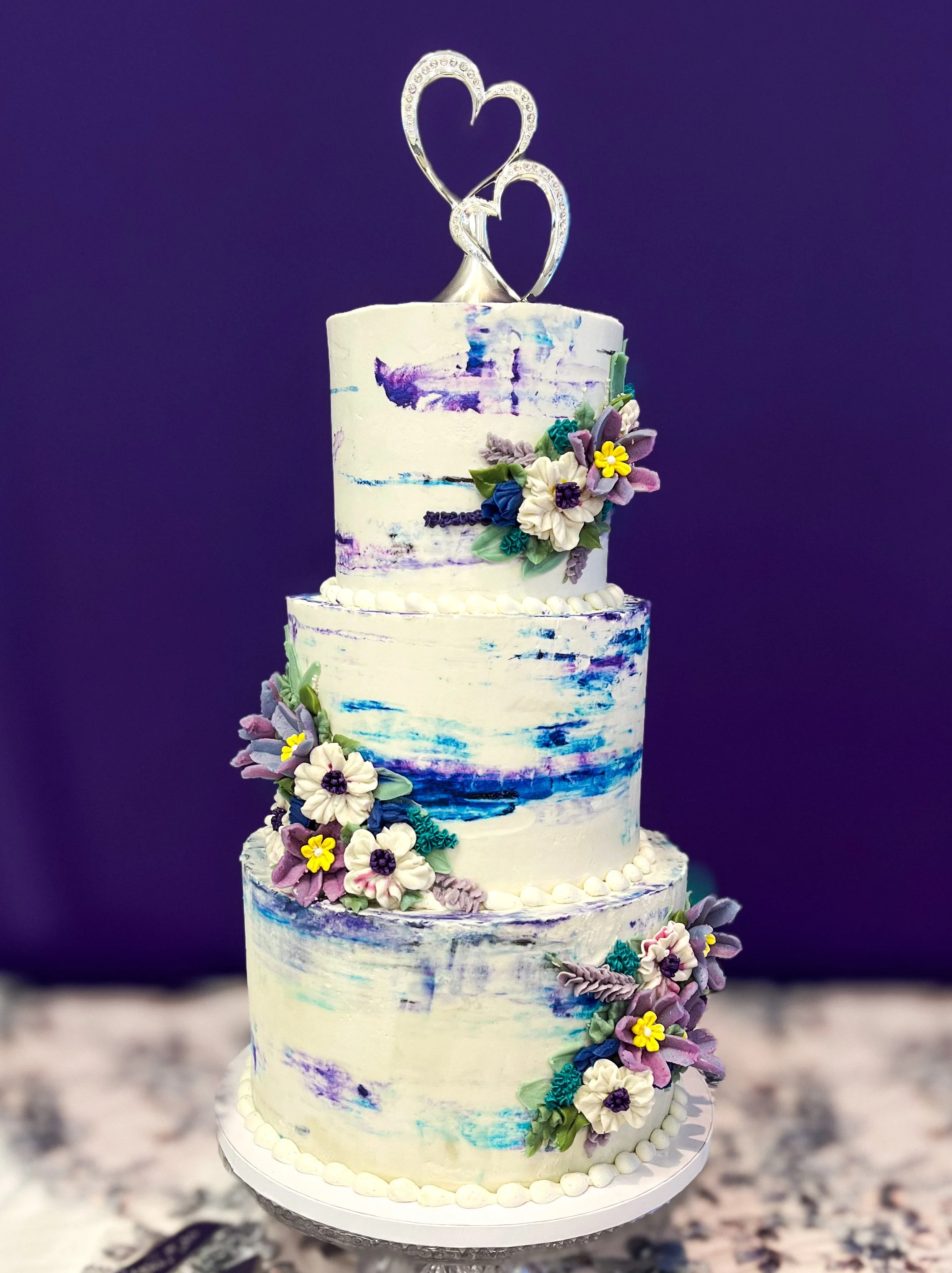 Three-tiered wedding cake with painted blue and purple strokes, decorated with colorful floral fondant, topped with heart-shaped silver cake toppers, on a white stand against a dark background.