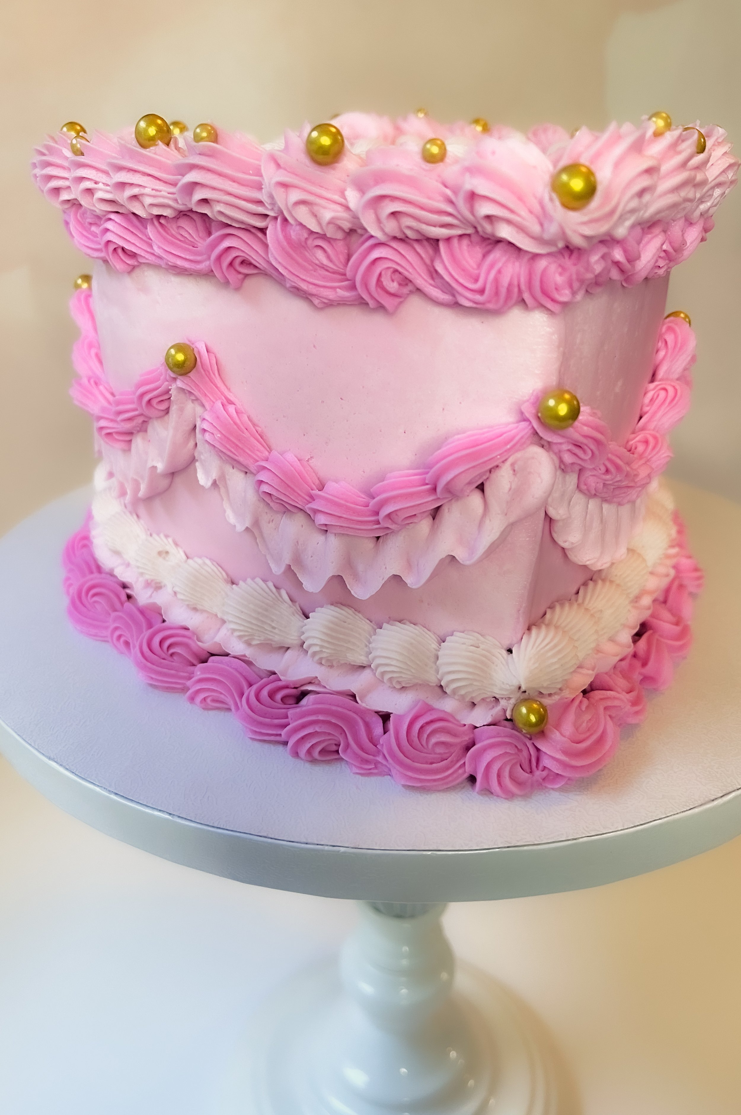 Two-tier pink cake decorated with pink and white frosting, gold edible beads, and piped decorations, placed on a white cake stand.
