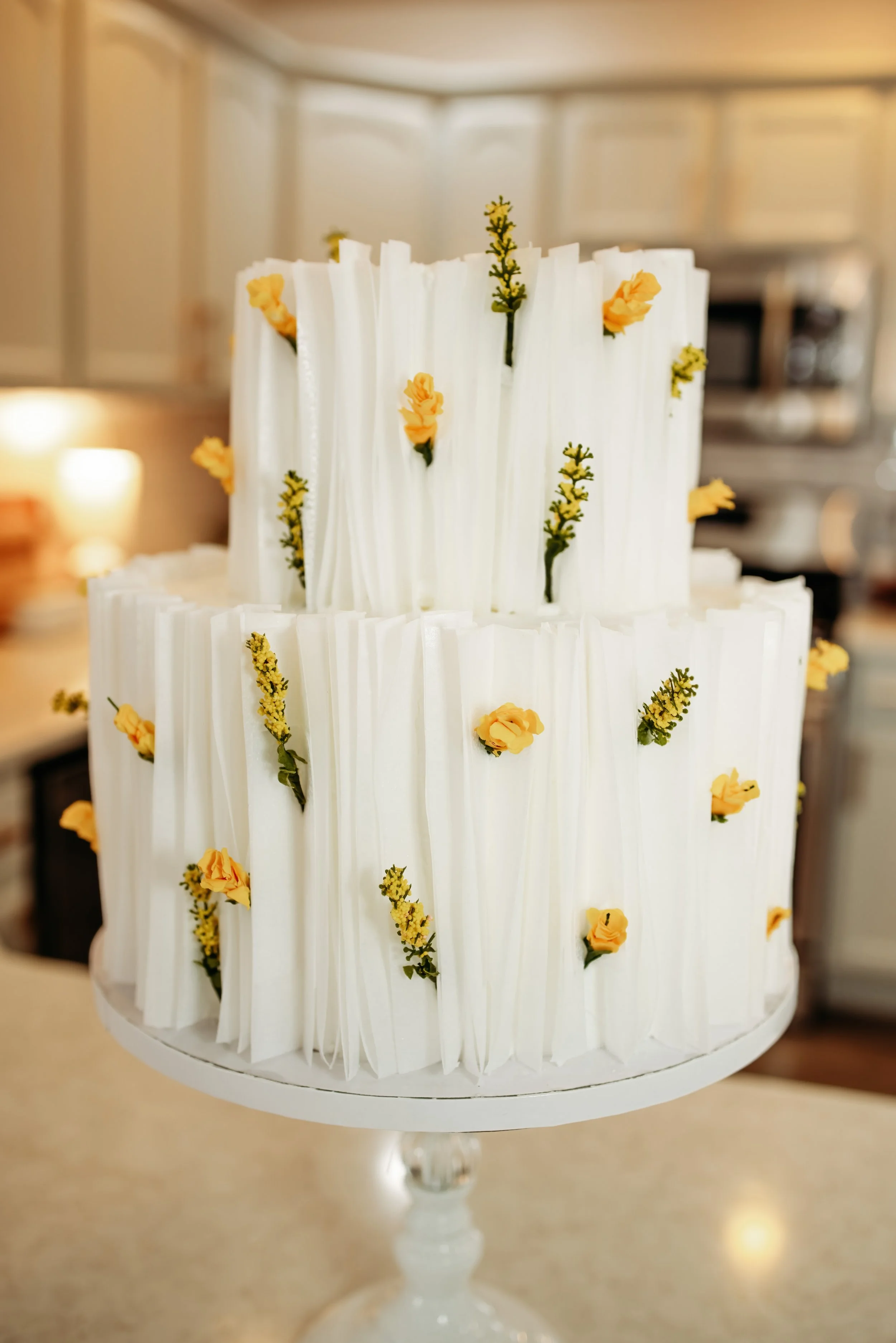 Two-tiered white wedding cake decorated with small yellow and green flowers, with vertical white fondant pleats on each tier, displayed on a white cake stand.