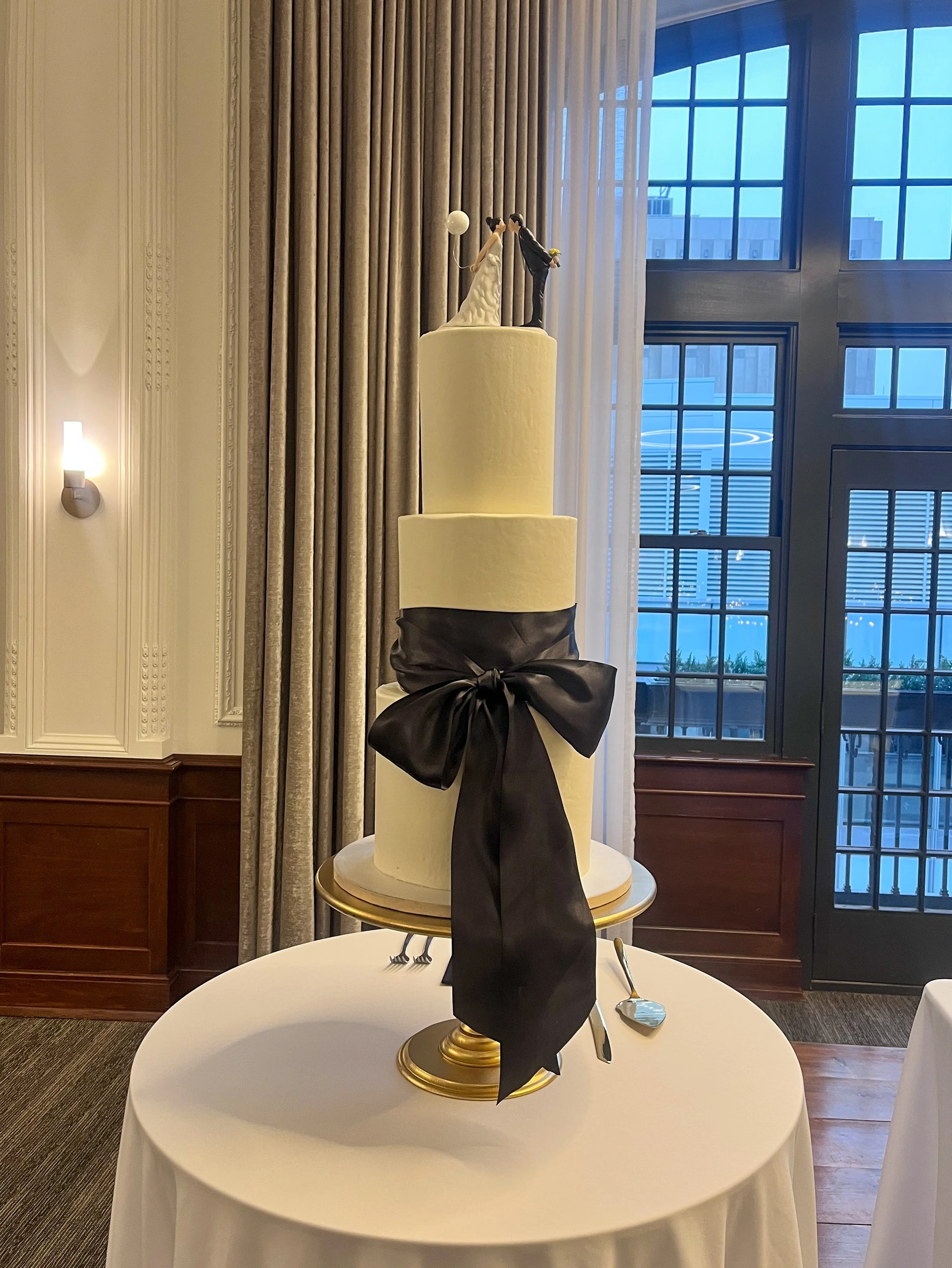Elegant wedding cake with two tall white tiers, decorated with a black ribbon bow, topped with a figurine of a bride and groom in wedding attire, set on a round table with silverware. The background features large windows and curtains.