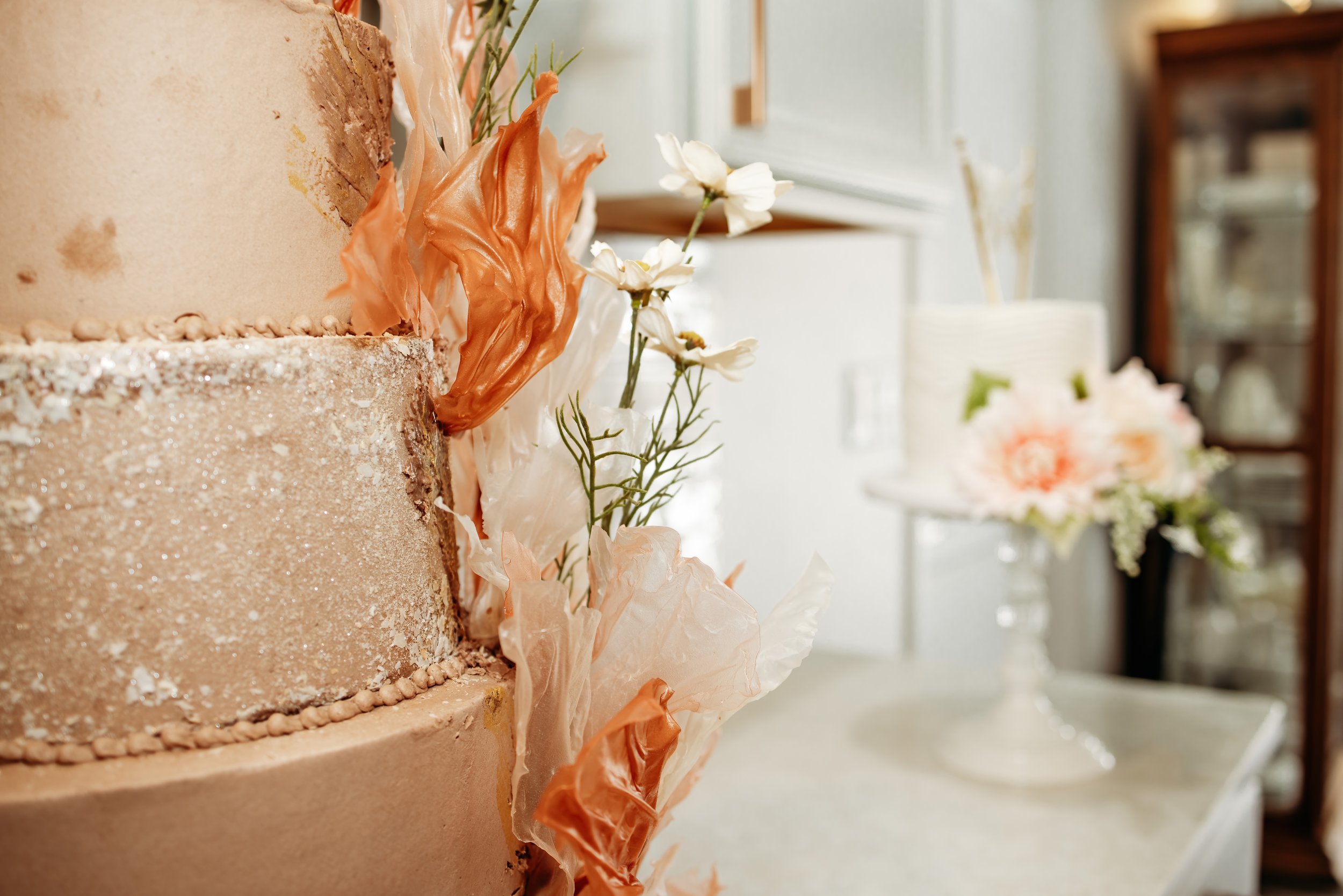 Close-up of a decorated beige wedding cake with peach-colored and white tissue paper flowers, with a blurred background showing a floral arrangement on a cake stand.