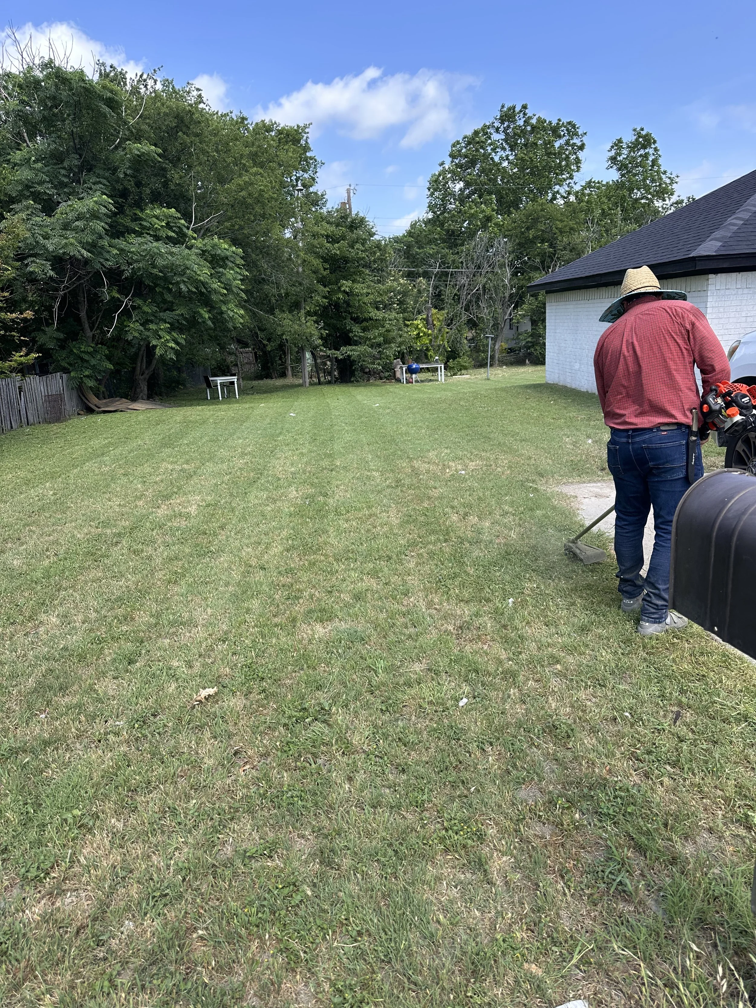 A man wearing a straw hat and red plaid shirt using a leaf blower in a backyard with grass, trees, a white building, and a clear blue sky.