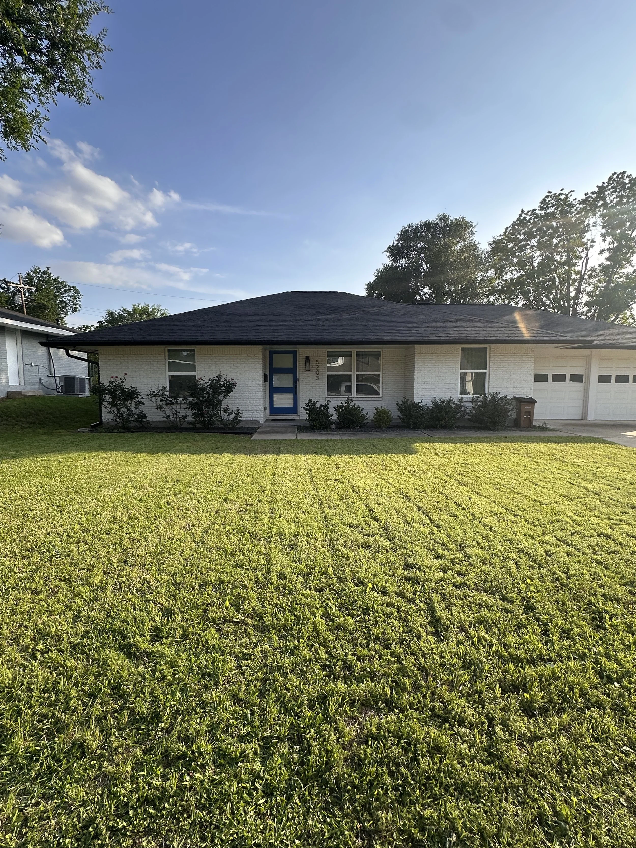 A single-story white house with a dark shingle roof, blue front door, and a garage, surrounded by a green lawn and bushes, with trees and a blue sky with clouds in the background.