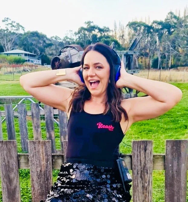 Woman with Silent Disco headphones on. sitting down with her hands over the headphones. she is wearing a black sequin skirt and a singlet top that says 'Off The Leash Tours'