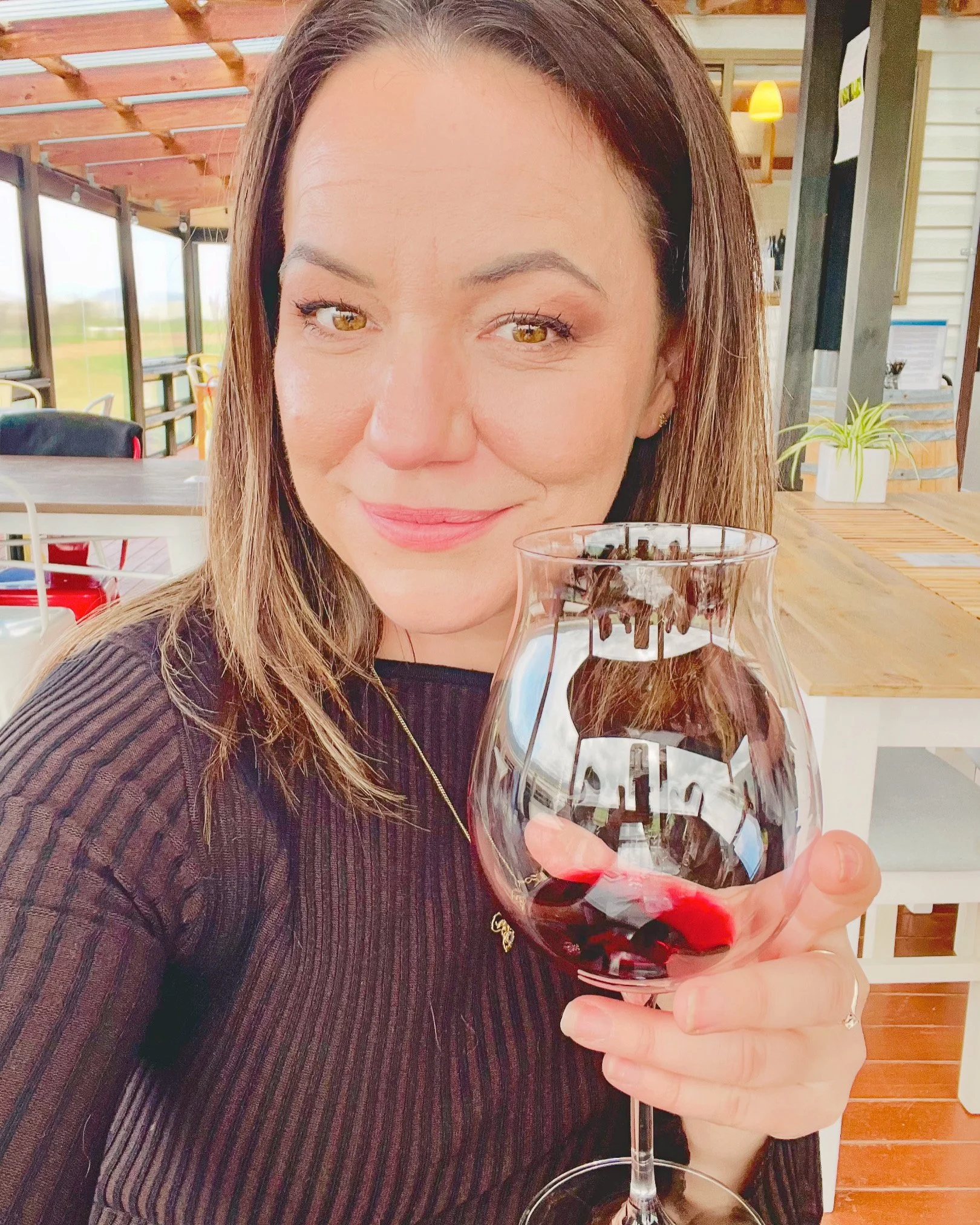 A woman with shoulder-length brown hair, wearing a black ribbed top, holds a glass of red wine, smiling and sitting at a wooden table in a bright, airy restaurant with large windows and wooden accents.