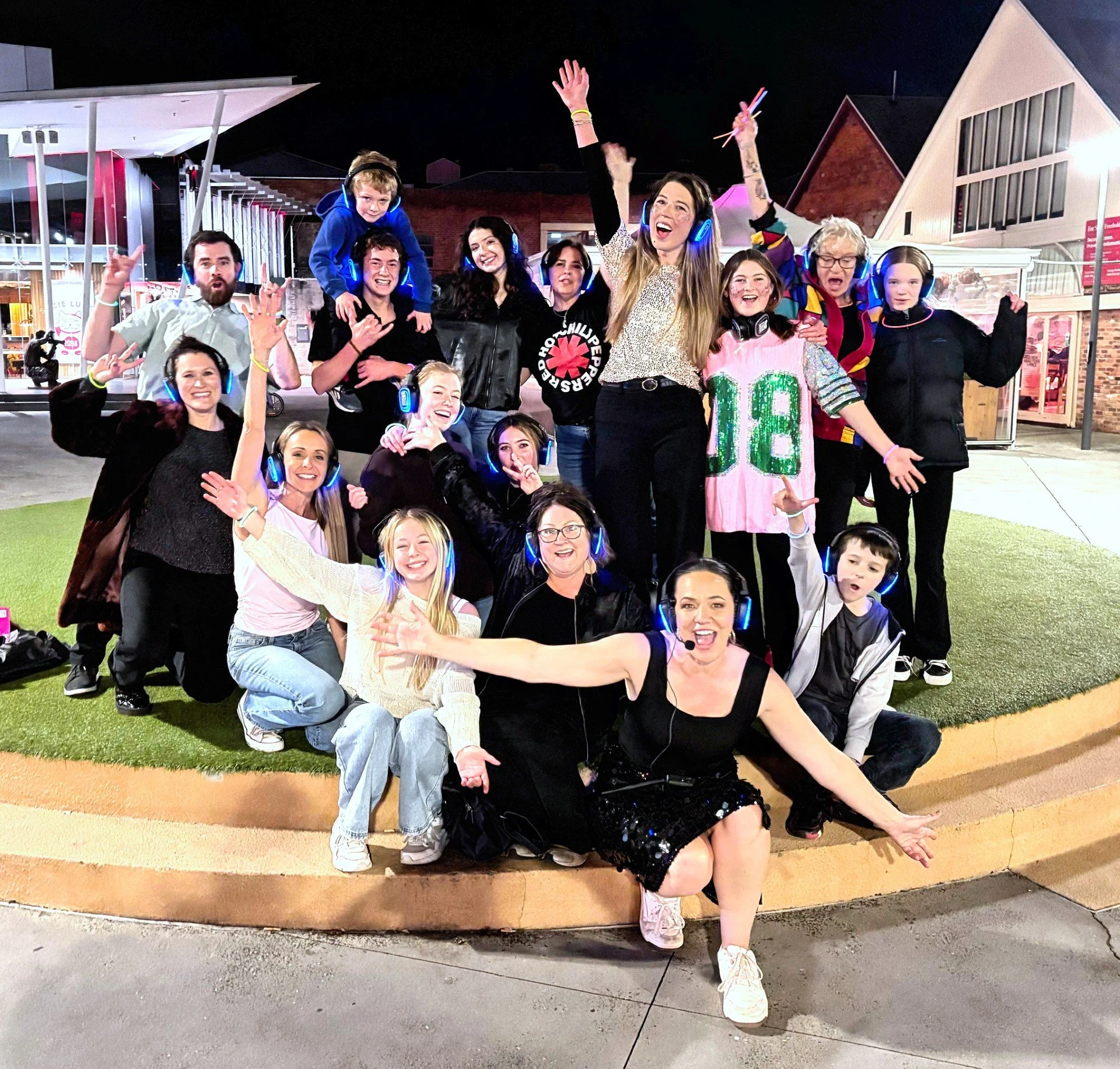 A group of diverse people, including children and adults, celebrating together at night outside. They are wearing headphones with lights, smiling, waving, and making various joyful gestures. The background features modern buildings and a dark sky.