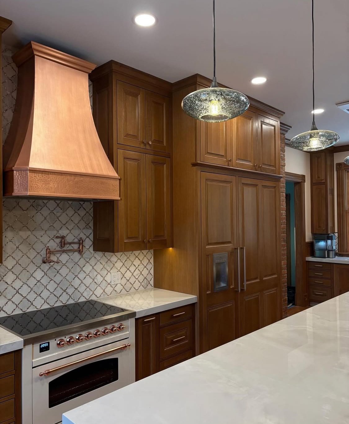 Kitchen with wooden cabinets, a copper range hood, a white oven with copper knobs, a patterned backsplash, and modern pendant lights.