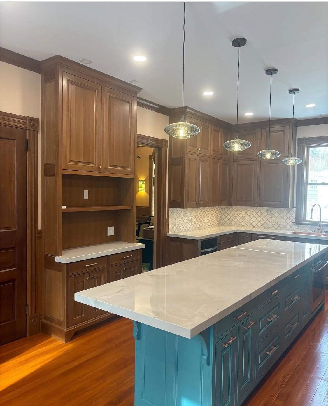 Custom remodeled kitchen with wooden cabinets, a large marble-topped island blue-painted base, pendant lights, and a tiled backsplash.