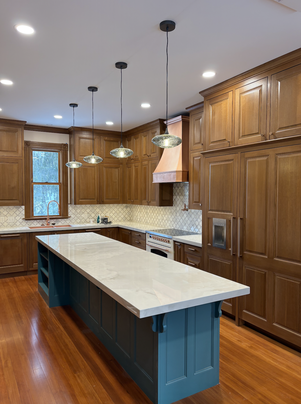 A spacious, custom kitchen that has been remodeled with wooden cabinets, white countertops, a large blue island, a copper hood over the stove, pendant lights, and hardwood flooring.