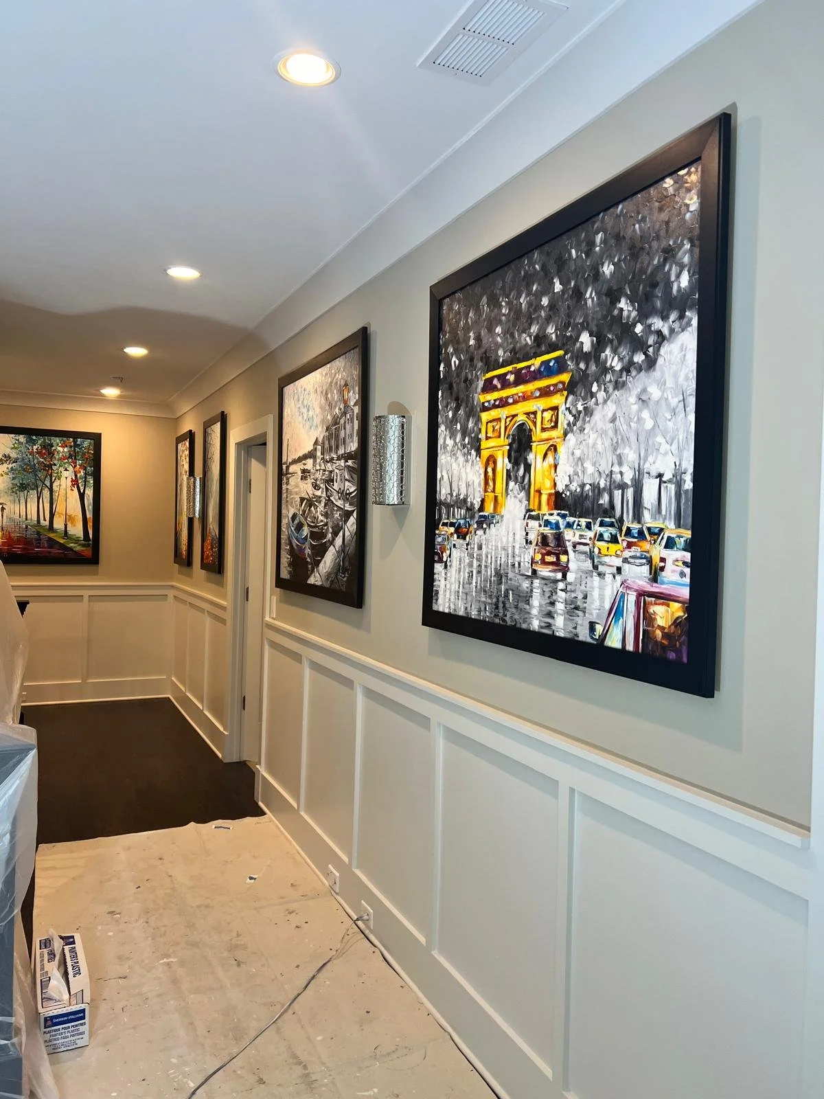 Hallway with beige walls, white wainscoting, ceiling lights, framed paintings, and a partially visible stairway. The floor appears to be under renovation.