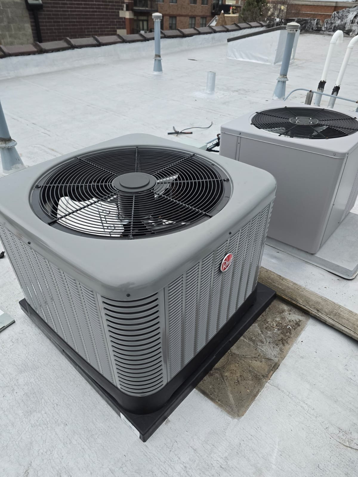 Rooftop HVAC units with a white flat roof, ventilation pipes, and a brick building in the background.