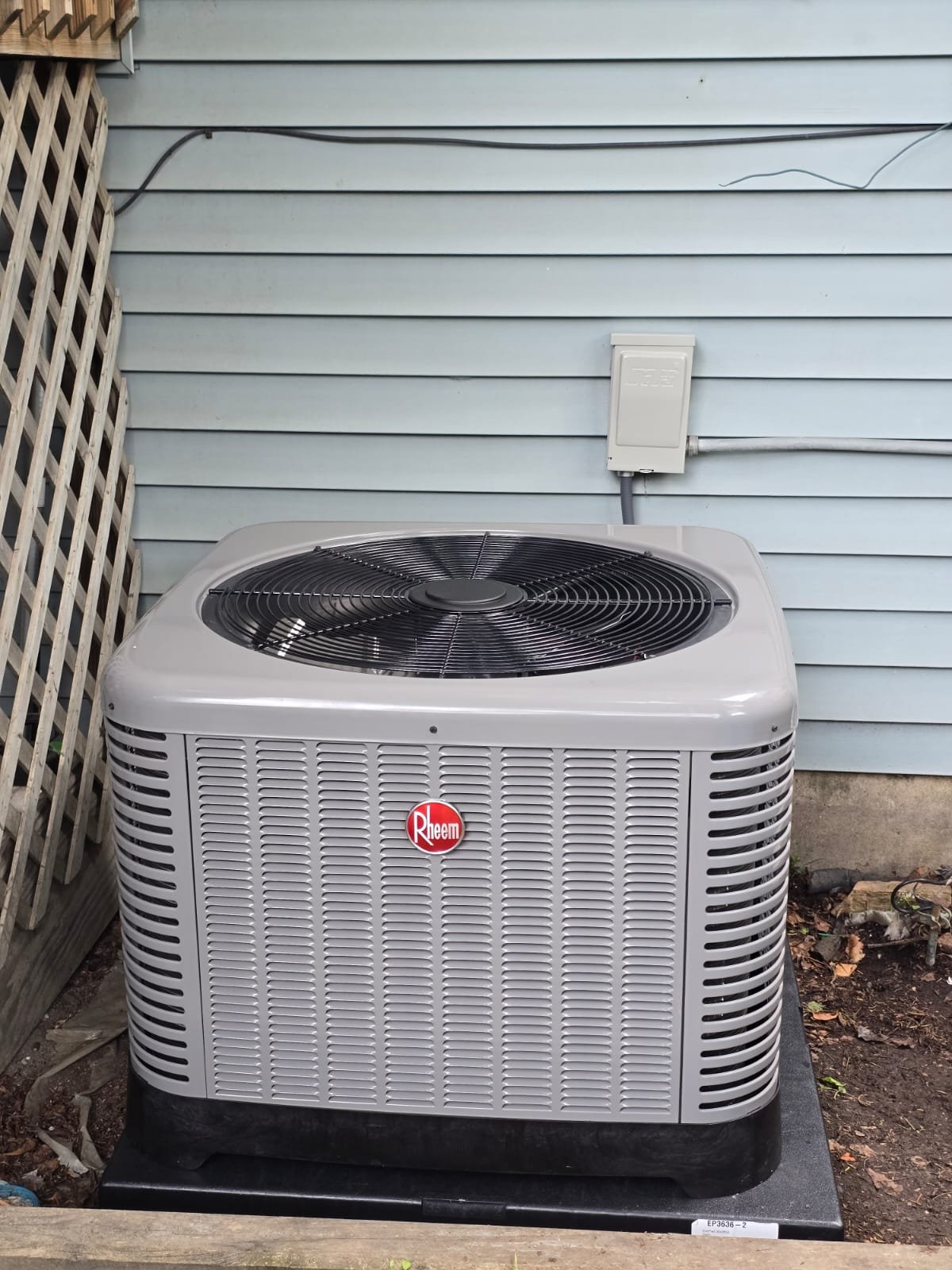 Outdoor Rheem air conditioning unit next to a light blue house with horizontal siding, a vented wooden lattice on the left, and an electrical box connected to the house's wall.