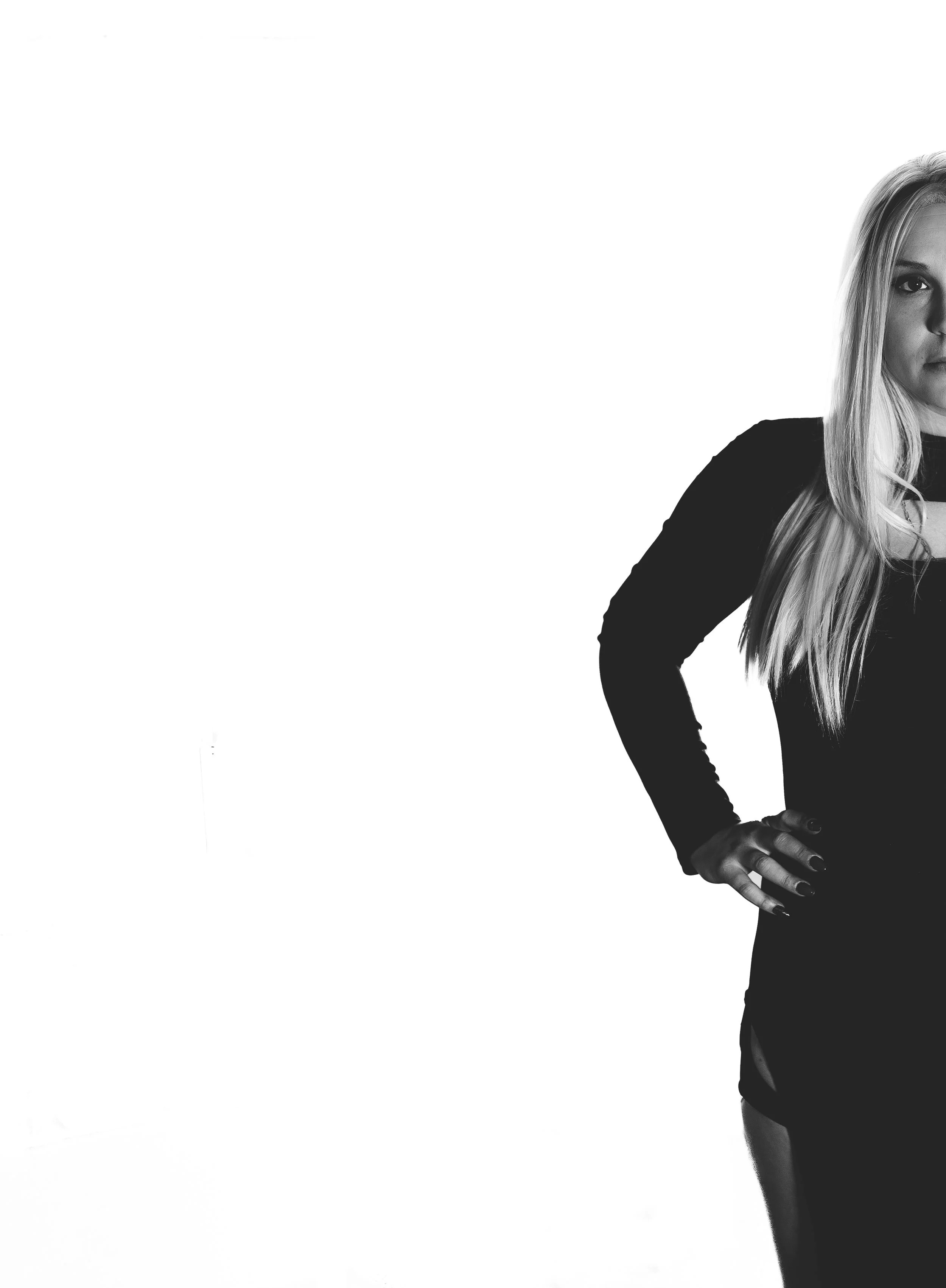 Black and white photo of a woman with long blonde hair, wearing a black long-sleeved top, with her left hand on her hip, against a white background.