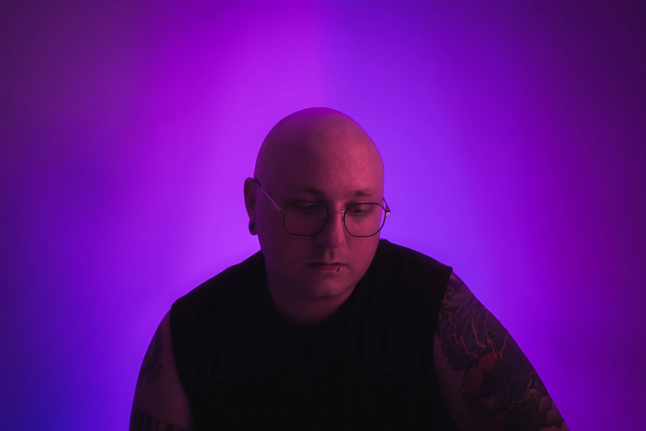 A person with a shaved head, glasses, and tattoos on their arms, looking down, illuminated by purple and blue lighting in a dark setting.