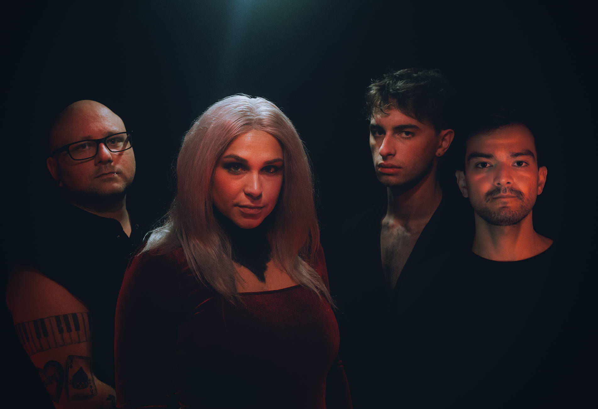 Four individuals posing in a dark studio with dramatic lighting, including a woman with long blonde hair and three men with varying hairstyles and facial expressions.