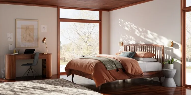A modern bedroom with a large window, a wooden bed with rust-colored bedding, a cozy blanket, and a small desk with a chair, lamp, and framed artwork.