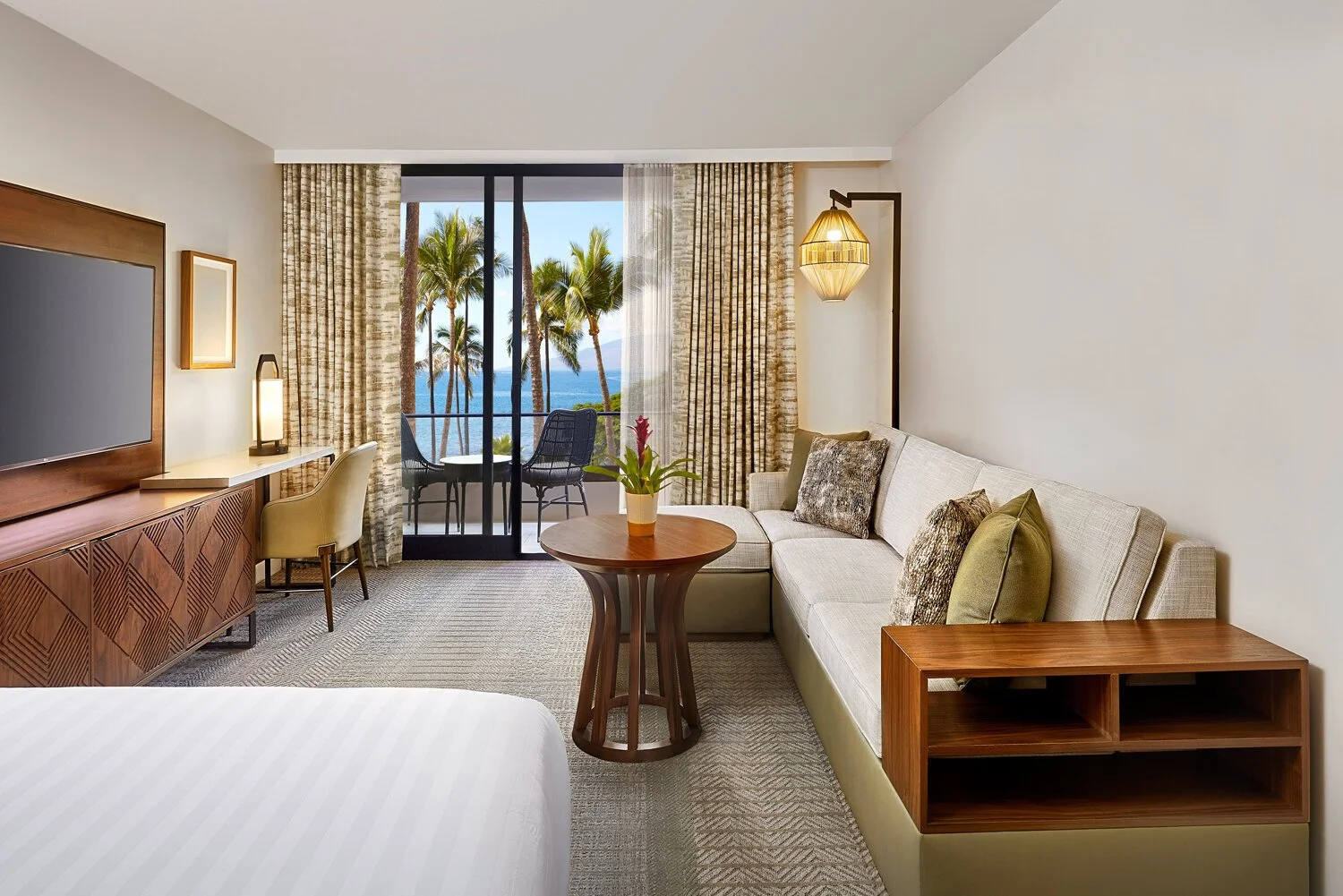 Luxury hotel room with a balcony overlooking palm trees and the ocean, featuring a cream sofa with pillows, a round wooden coffee table with a potted plant, a flat-screen TV, a desk with a chair, and large curtains.