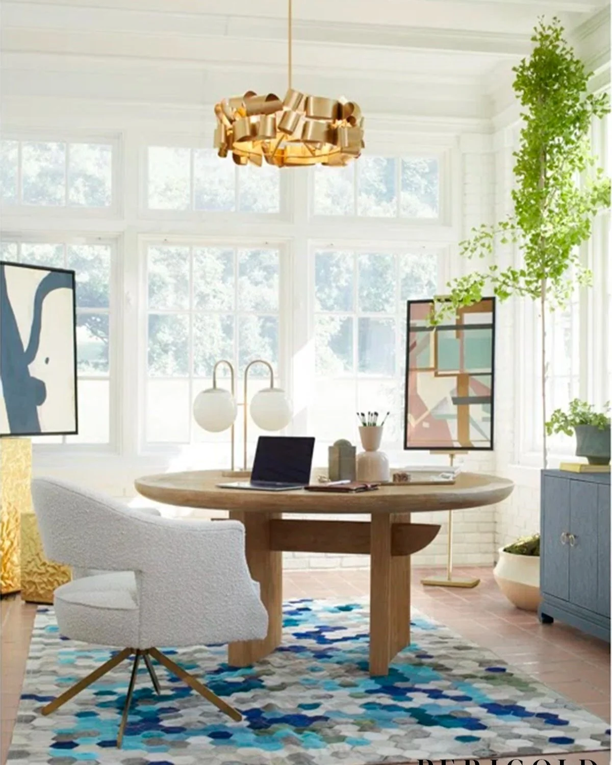 Bright dining room with large windows, modern chandelier, round wooden table, white upholstered chair, colorful geometric rug, blue sideboard, and abstract wall art, with green plants.