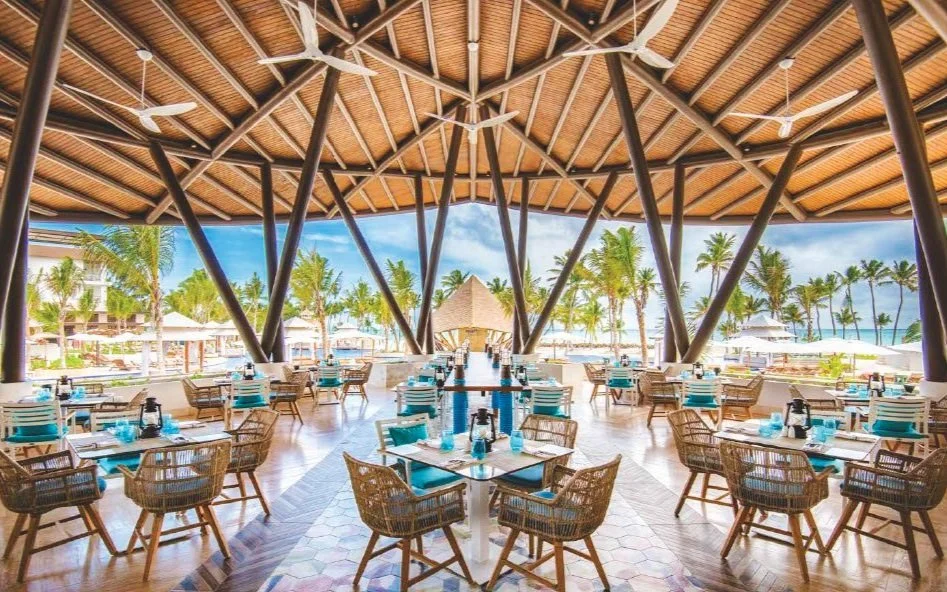 Open-air restaurant with wooden ceiling and large windows revealing a beach with palm trees. Multiple tables set with turquoise napkins and lanterns, surrounded by wicker chairs.