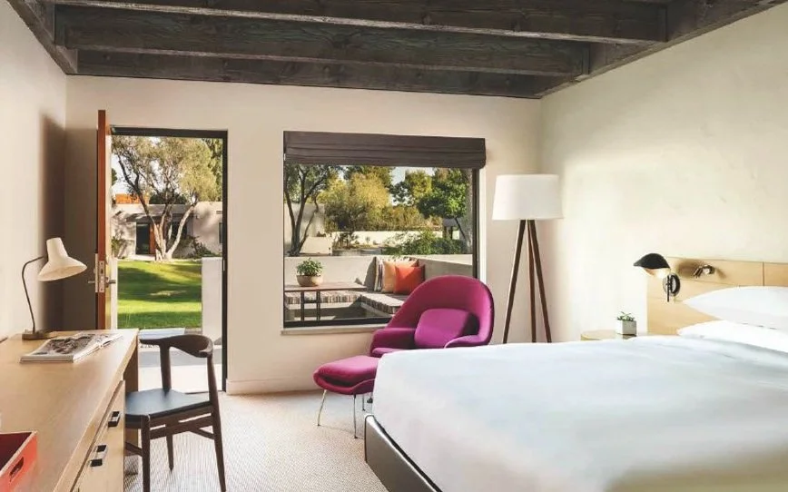 A modern hotel room with a bed, a pink lounge chair, a floor lamp, a desk with a chair, and a window with an outdoor view of trees and a patio.