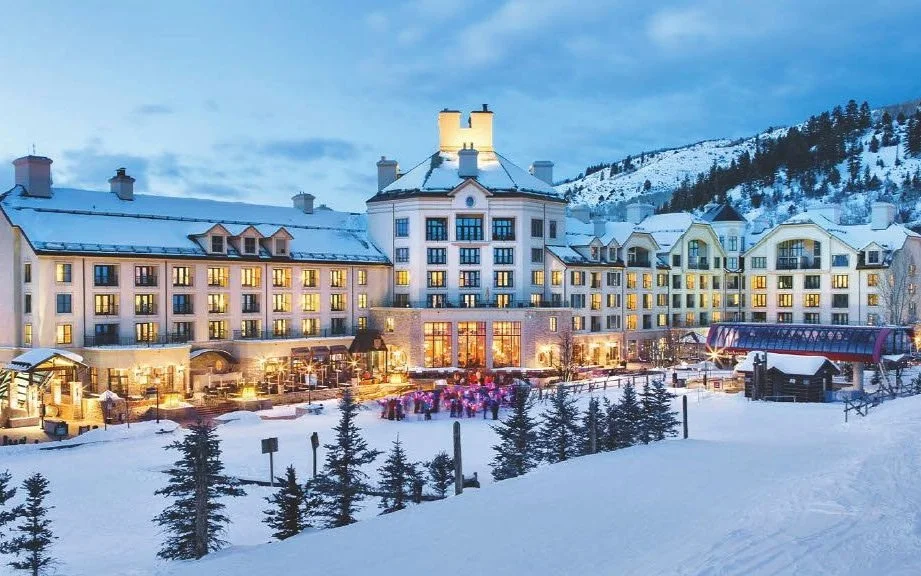 Snow-covered hotel with lit windows, surrounded by pine trees and mountains, during evening.