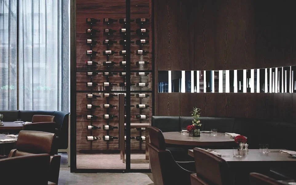 Interior of a restaurant featuring a wooden wine rack, seated tables with floral centerpieces, black leather chairs, and a large window with curtains.
