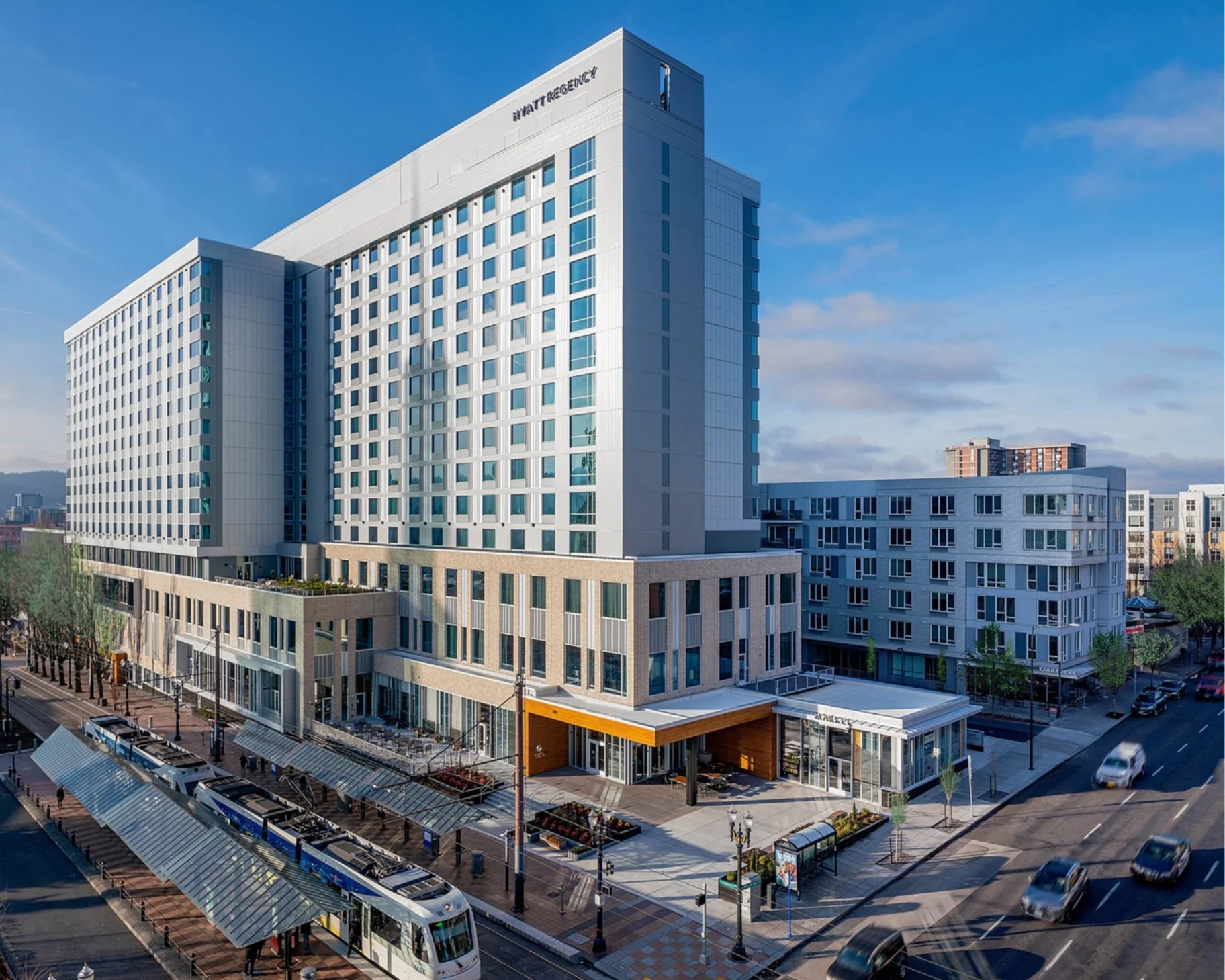 Modern multi-story hotel named 'Hilton' with a light-colored facade, situated on a city street with a streetcar and cars, trees, and a clear blue sky.