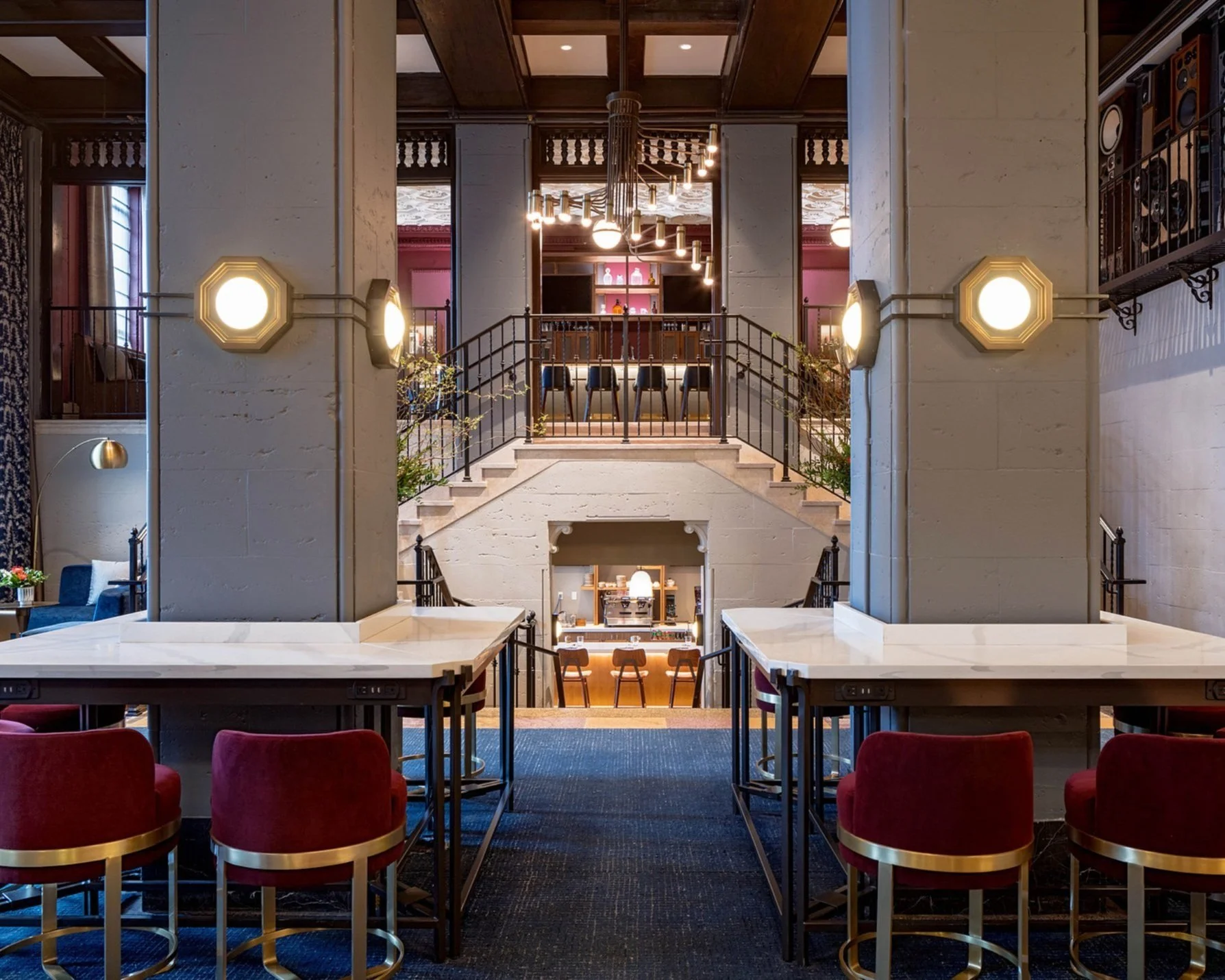 Interior view of a stylish, multi-level restaurant or cafe with marble-topped tables, red velvet chairs, decorative lighting, and staircase leading to an upper level with bar seating and artwork on the walls.