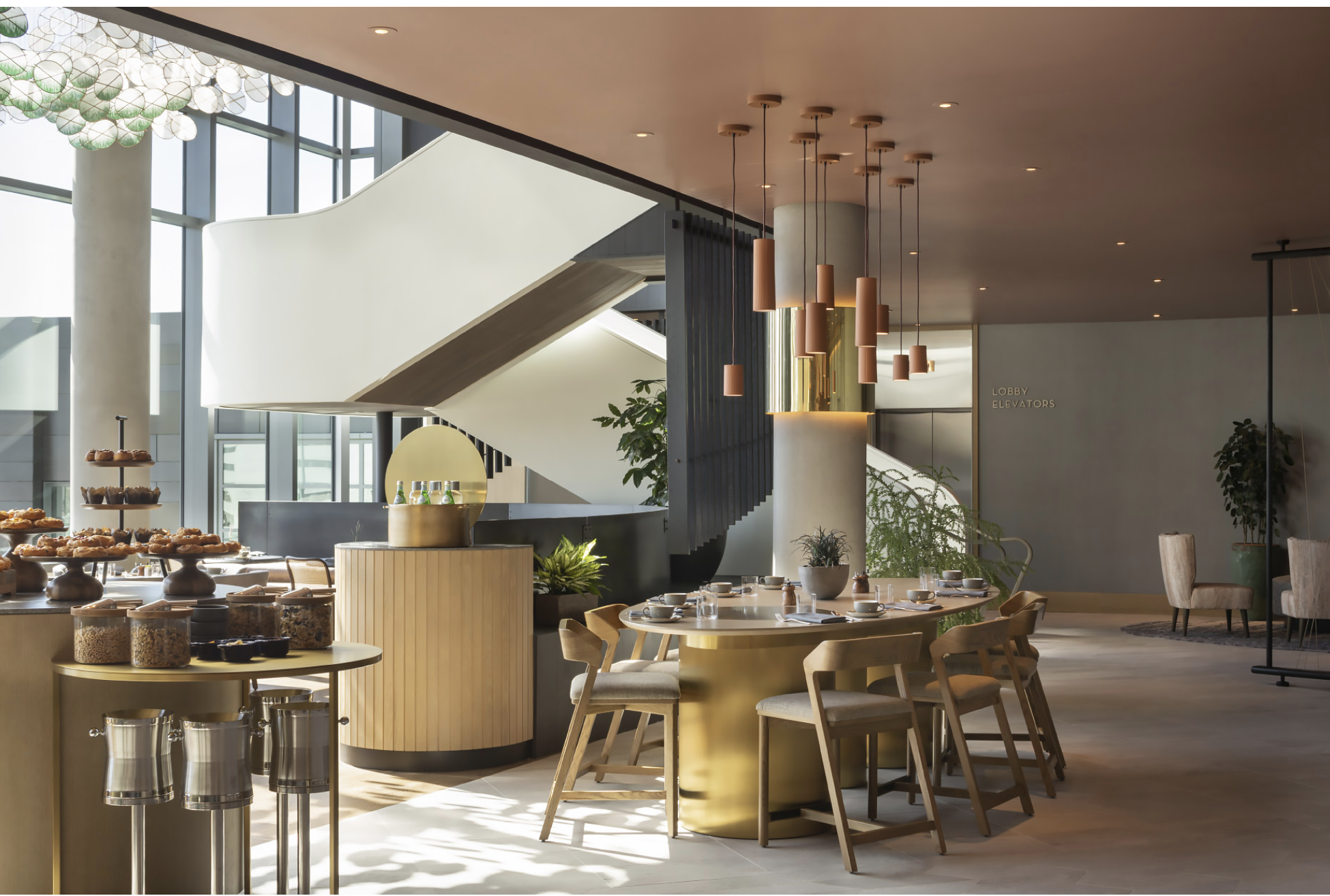 Modern dining area with a round table set with plates and glasses, surrounded by beige chairs, and a breakfast buffet with pastries and jars of cereal on a gold and white counter. Large windows and a spiral staircase are visible in the background.